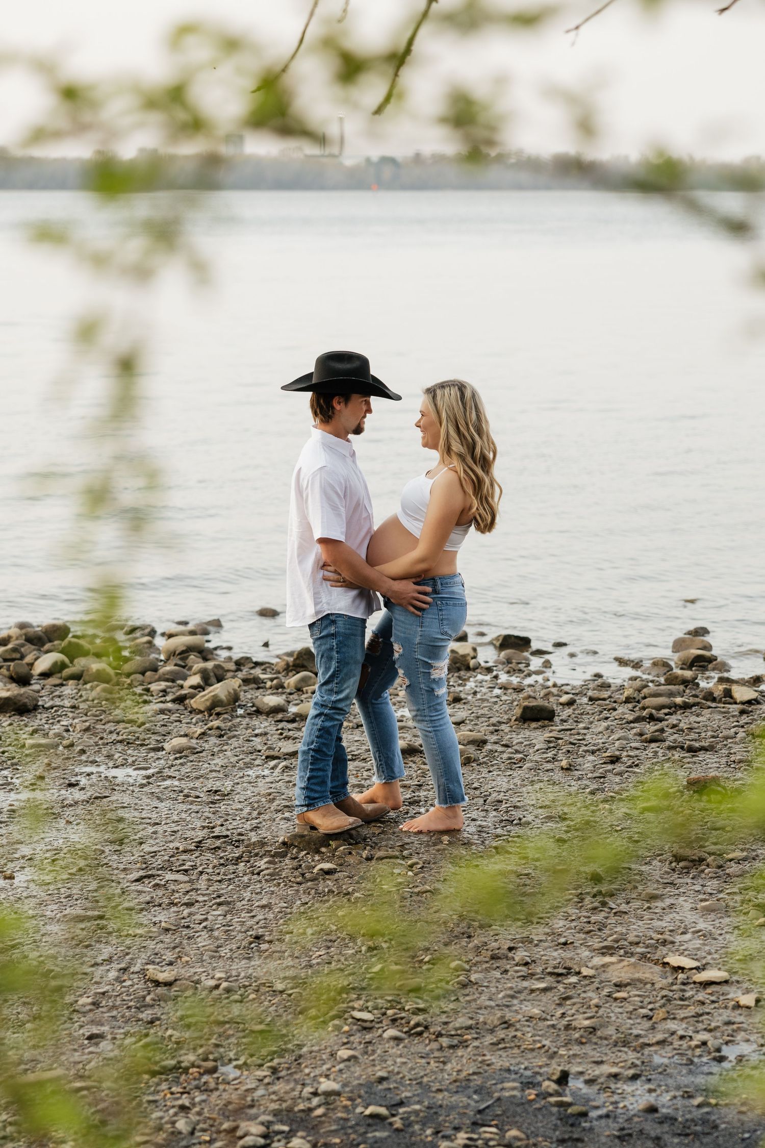 Pregnant couple in matching jeans and white tops smiling at each other on rocky riverside shore.