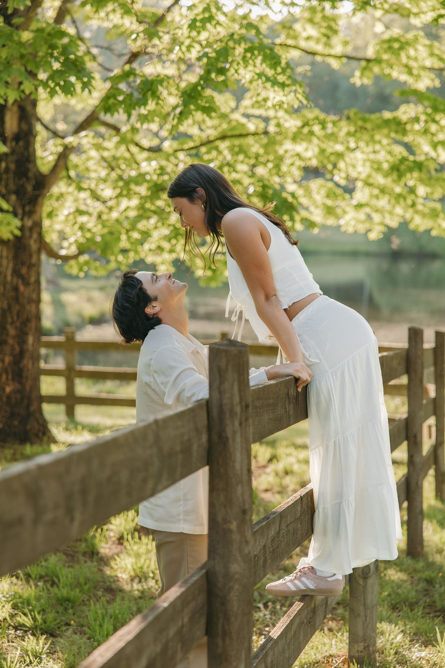 Couple gazing at each other with woman perched on a rustic wooden fence in a sunny garden.
