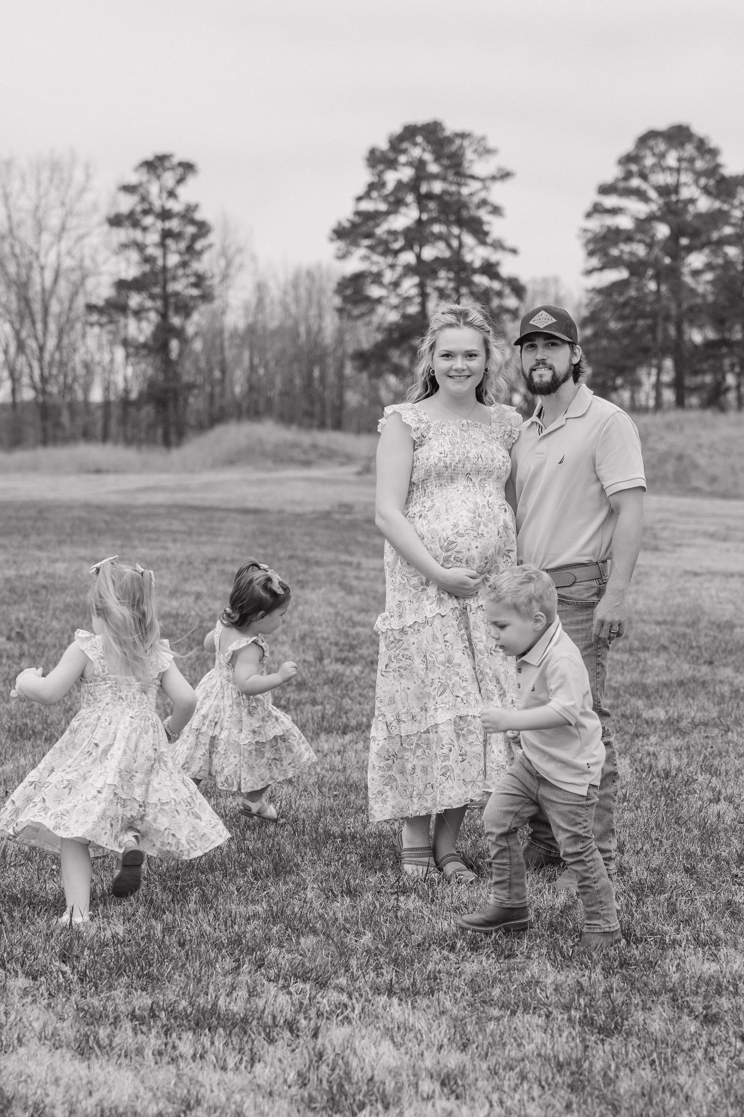 Black and white photo of pregnant woman and bearded man with three young children playing around them in grassy field.