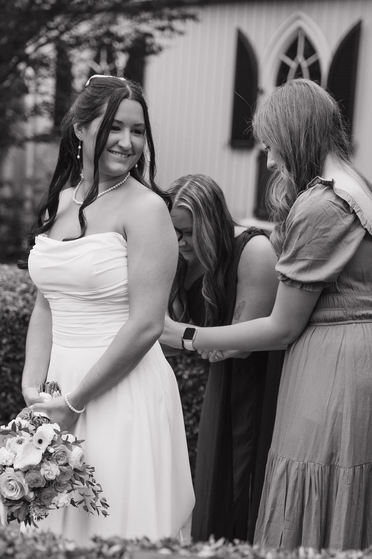Bride smiling in strapless white gown holding bouquet while bridesmaids adjust her dress outside a church.