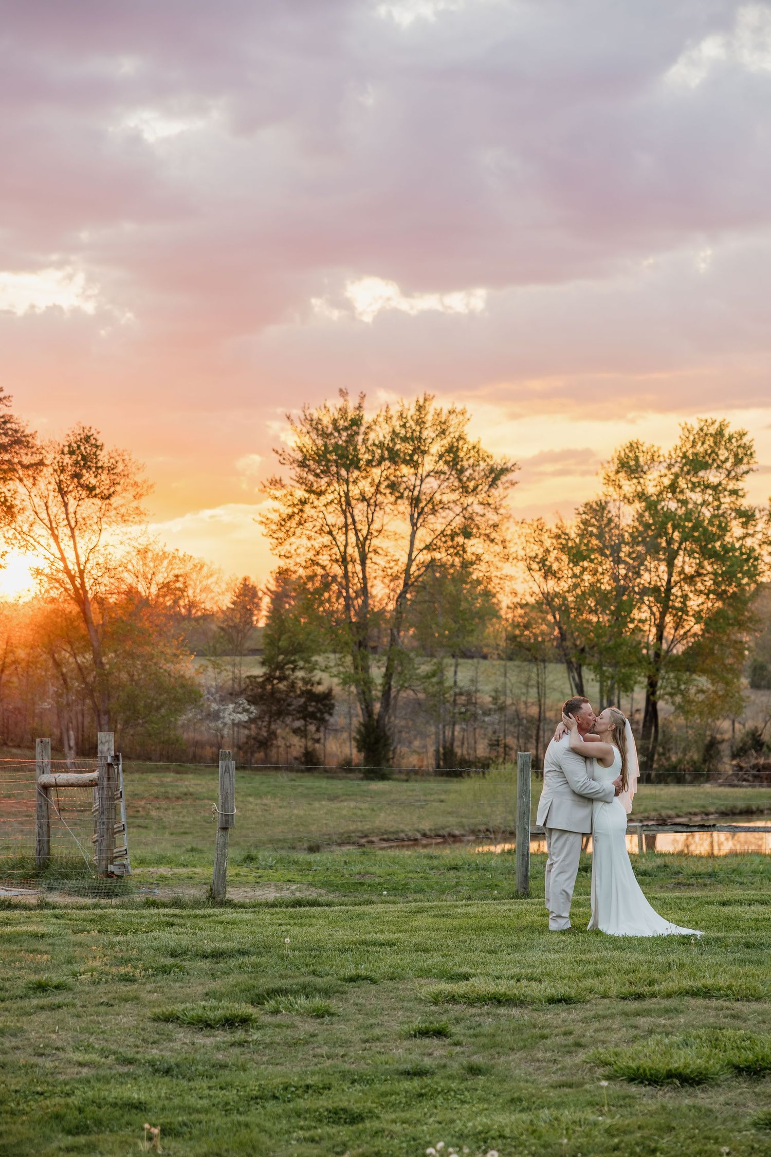 Newlyweds embracing near rustic fence posts with a stunning orange and pink sunset backdrop.