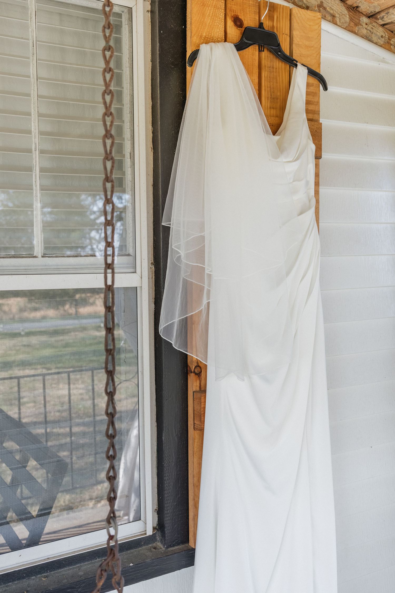 White wedding dress with veil hanging on wooden door of rustic farmhouse porch.
