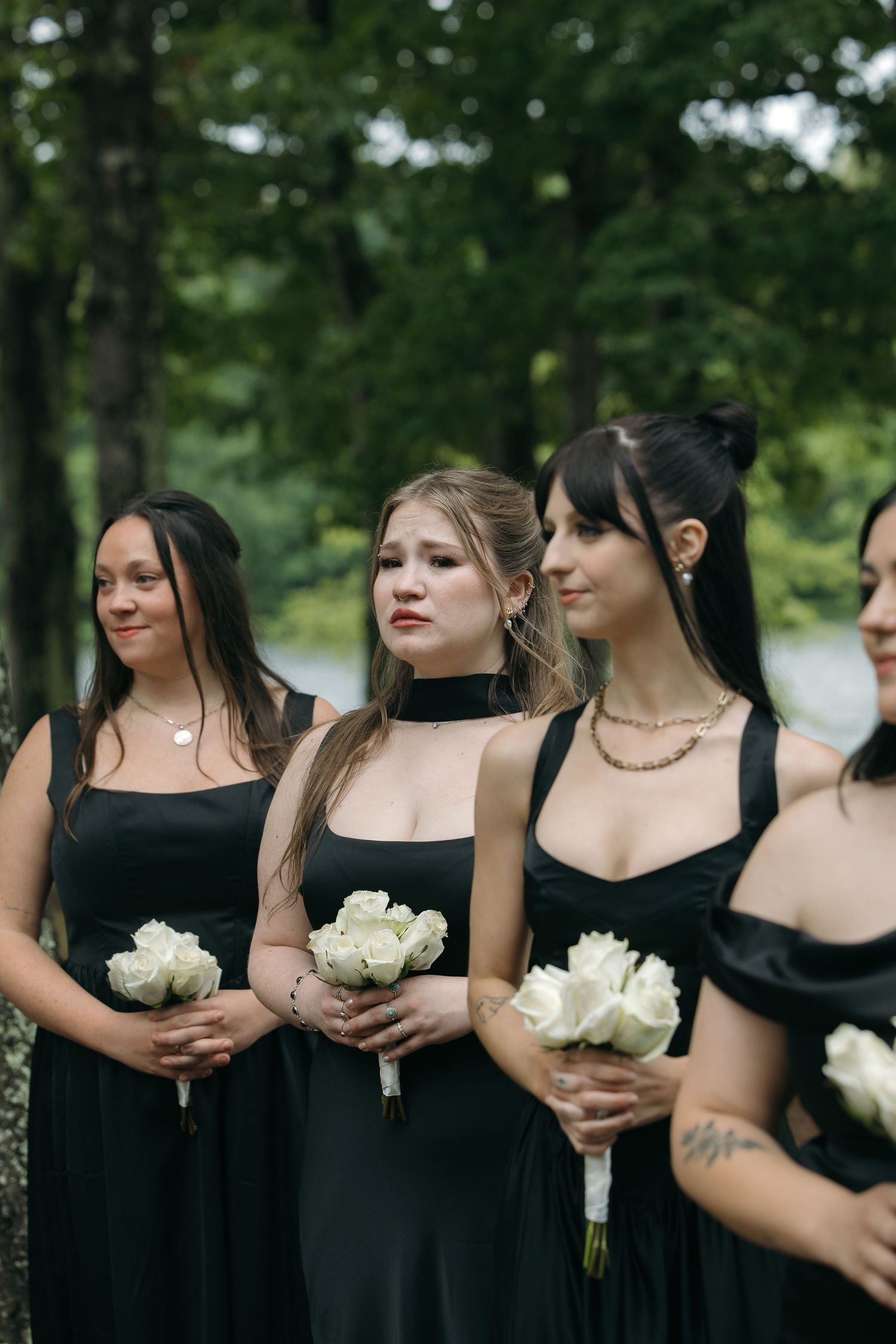 Four bridesmaids in black dresses holding white rose bouquets standing outdoors at wedding ceremony.