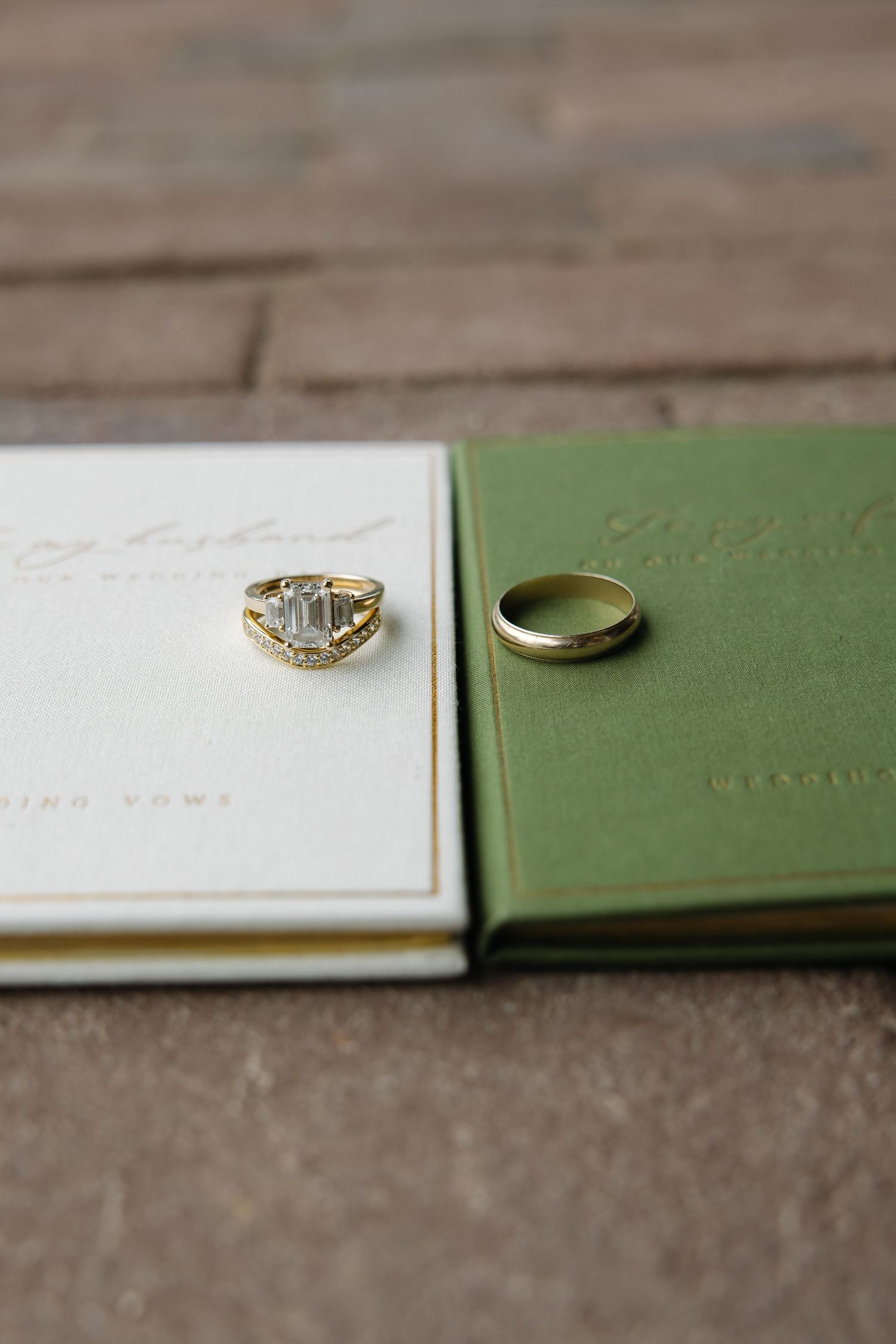 Gold wedding band and diamond engagement ring resting on white and green vow books on a stone surface.