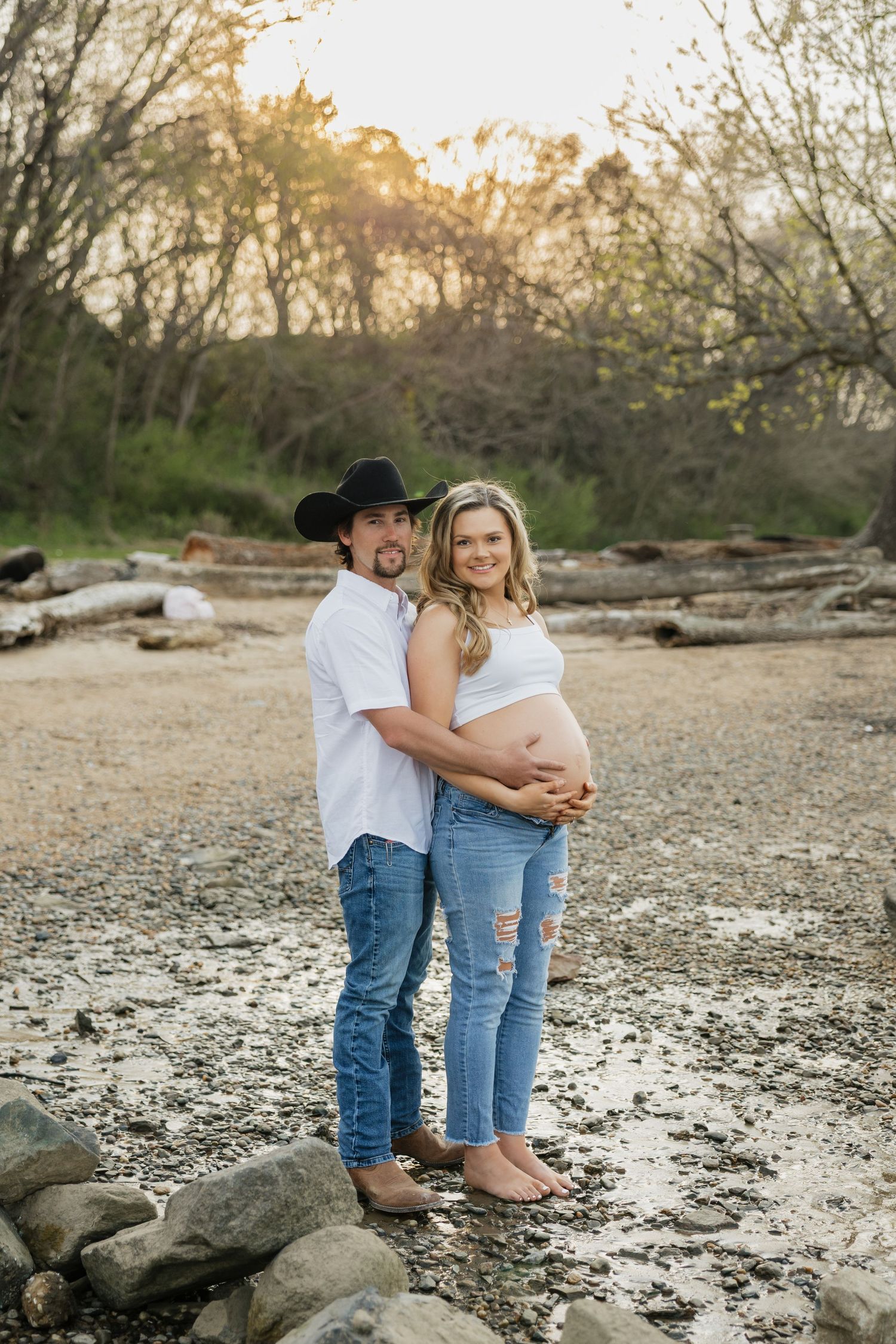 Couple poses for maternity photos at sunset on rocky shore, man in cowboy hat embracing pregnant woman in white crop top.