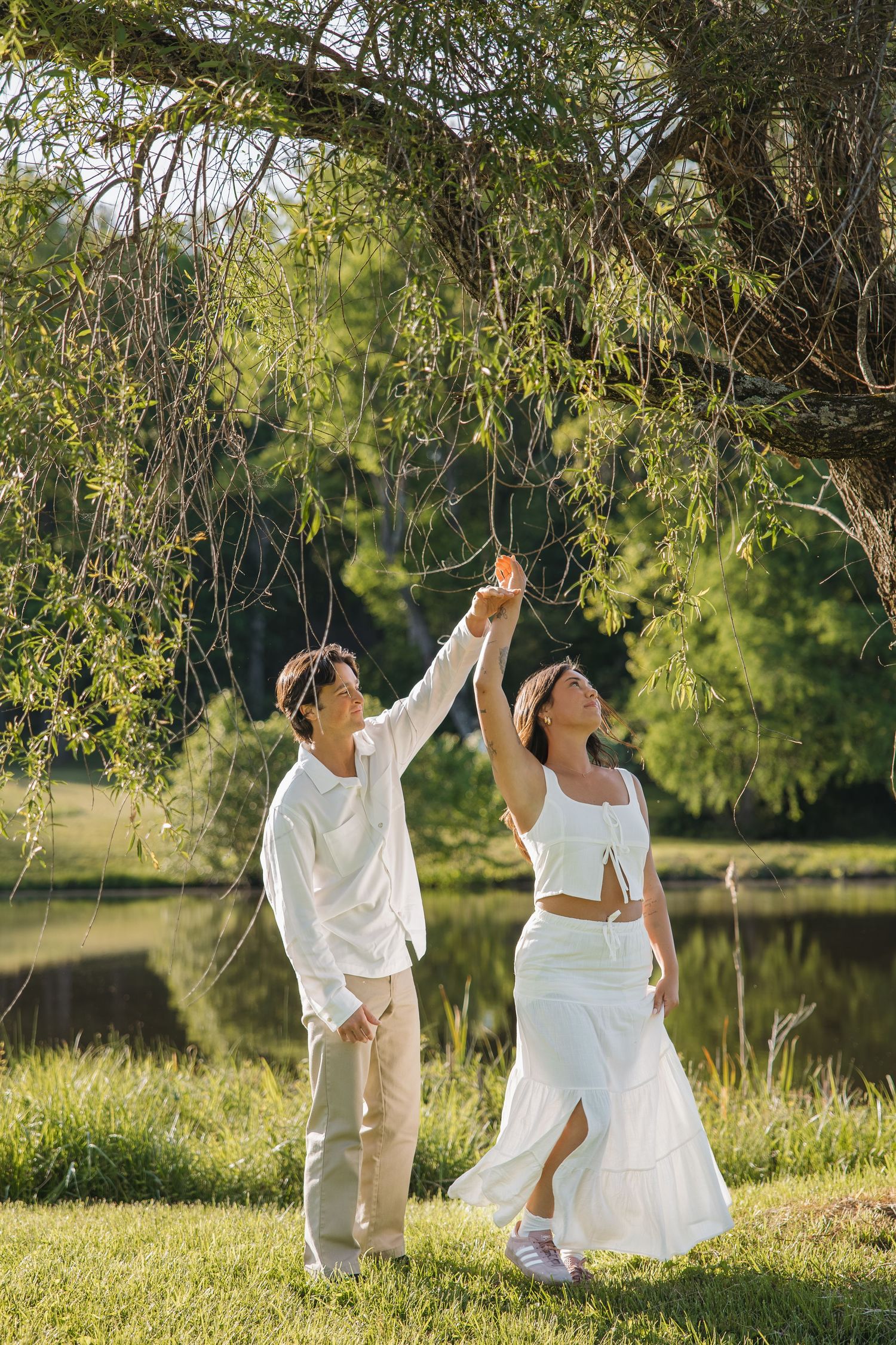 Couple in white outfits reaching up to touch willow tree branches beside a serene pond in golden sunlight.