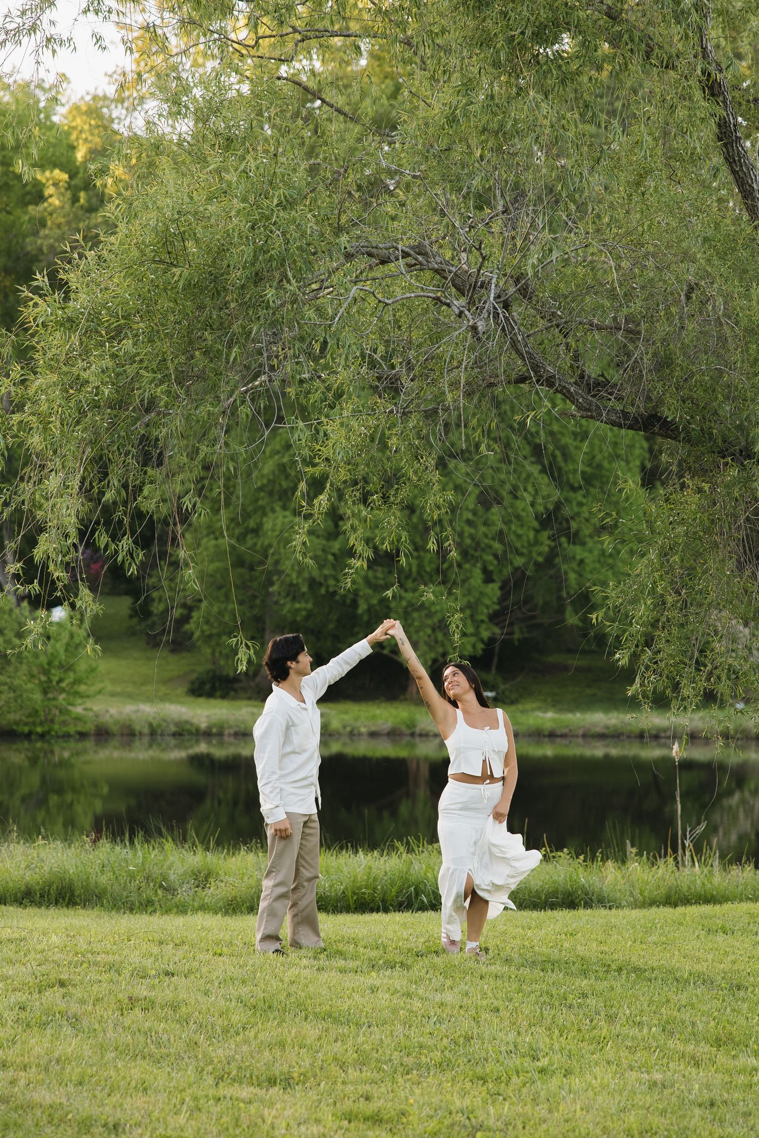 Couple twirling and dancing joyfully on green grass near a willow tree and peaceful pond.