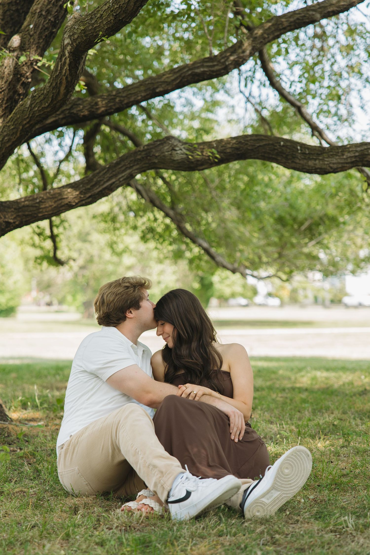 A couple sits closely together on grass beneath a large tree, sharing an intimate moment in a park.
