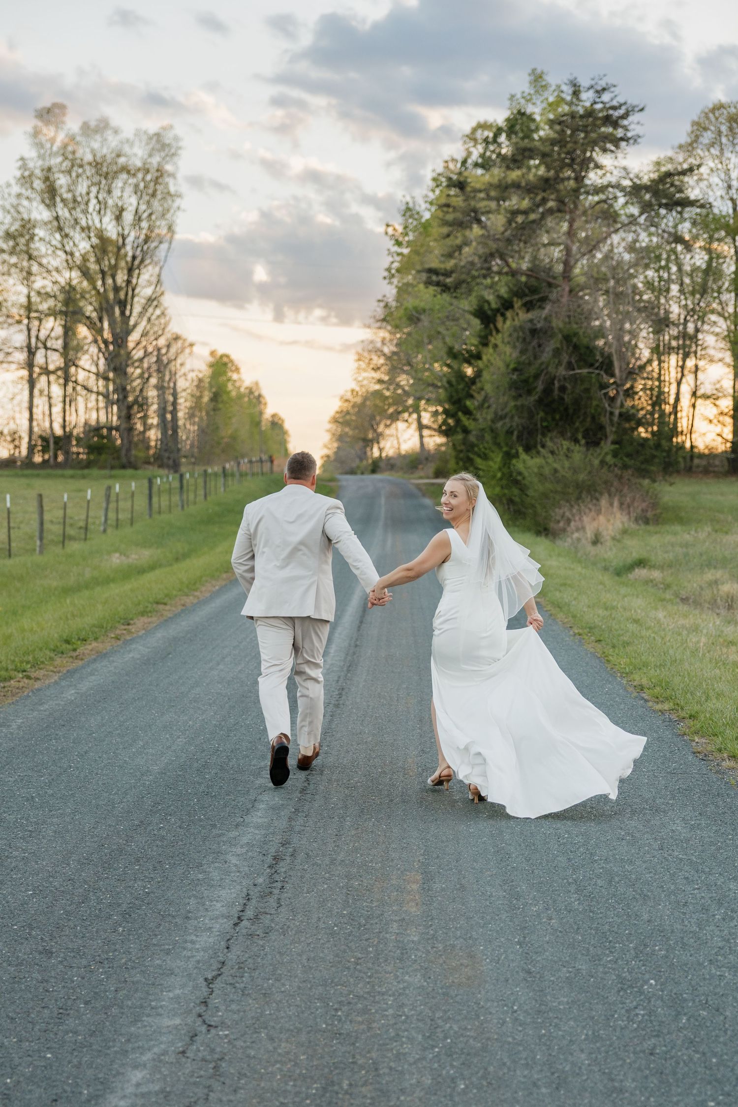 Bride looks back smiling as couple walks down a tree-lined road at sunset.