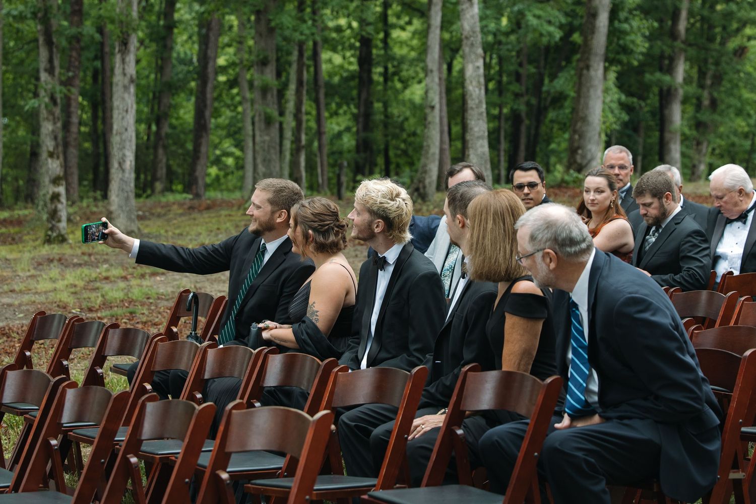 Wedding guests seated in wooden chairs taking a selfie at an outdoor forest ceremony.