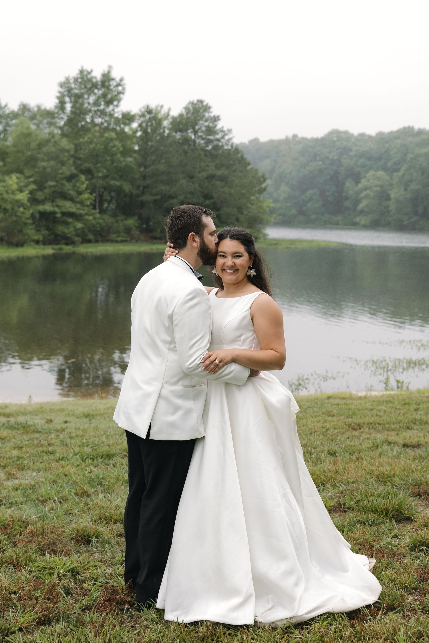 Groom in white jacket kissing bride's cheek as she smiles by a serene tree-lined lake.