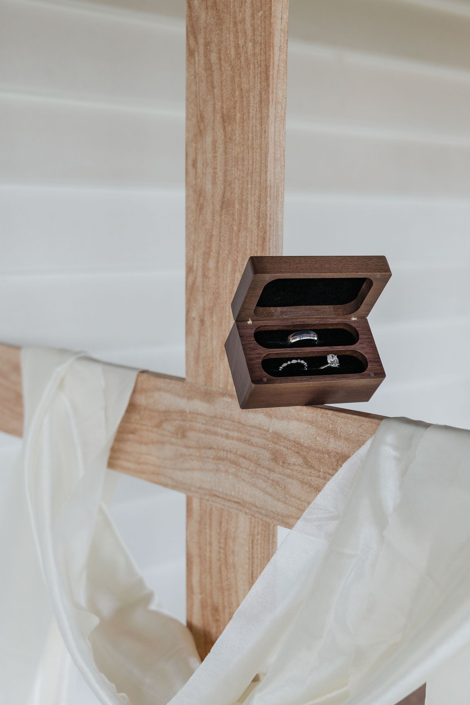 Elegant wooden ring box engraved with Ephesians 5:31 scripture displayed on wooden cross with white draping at wedding.