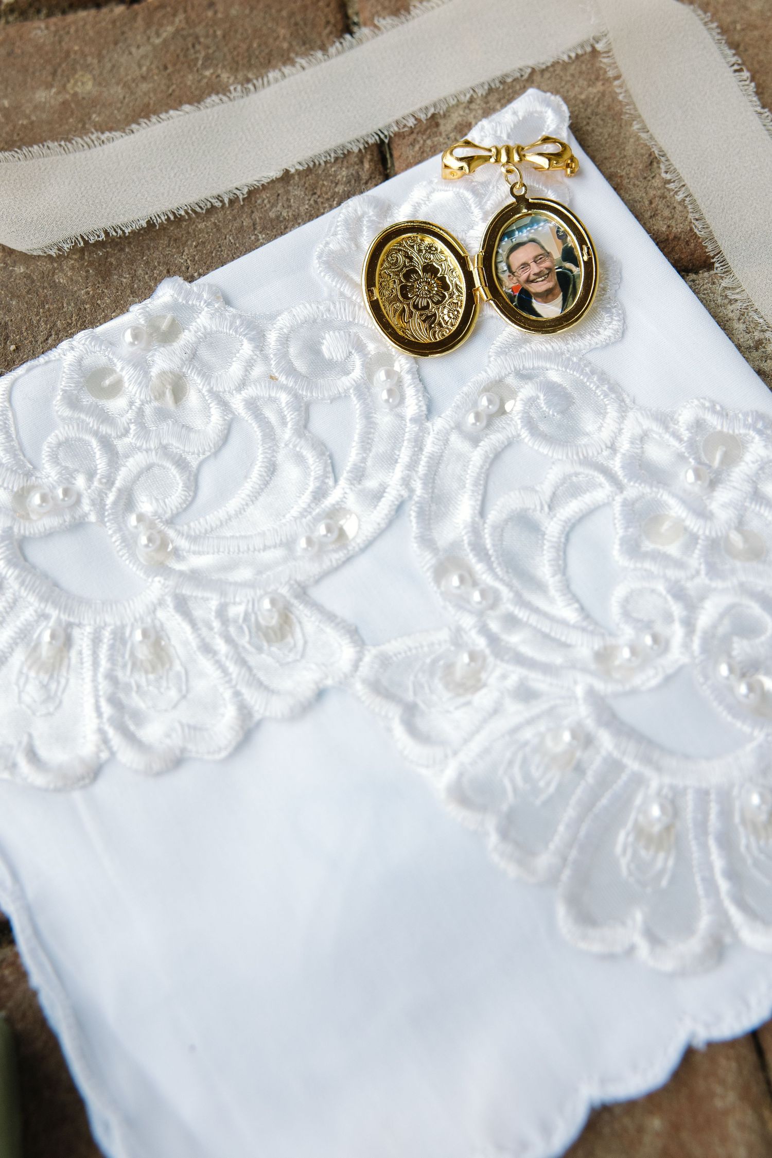 Lace embroidered white wedding handkerchief with gold locket photo charms displayed on brick surface.