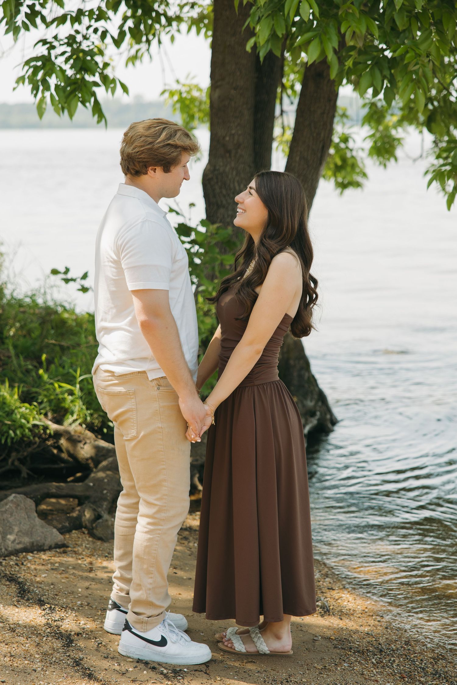 Young couple holding hands and gazing at each other by a lakeside tree on a sunny day.