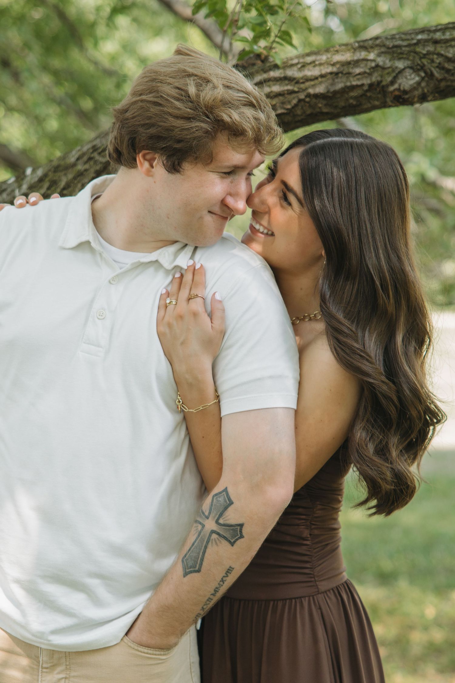 A couple gazes lovingly at each other under tree branches, woman in brown dress with cross tattoo embracing man.