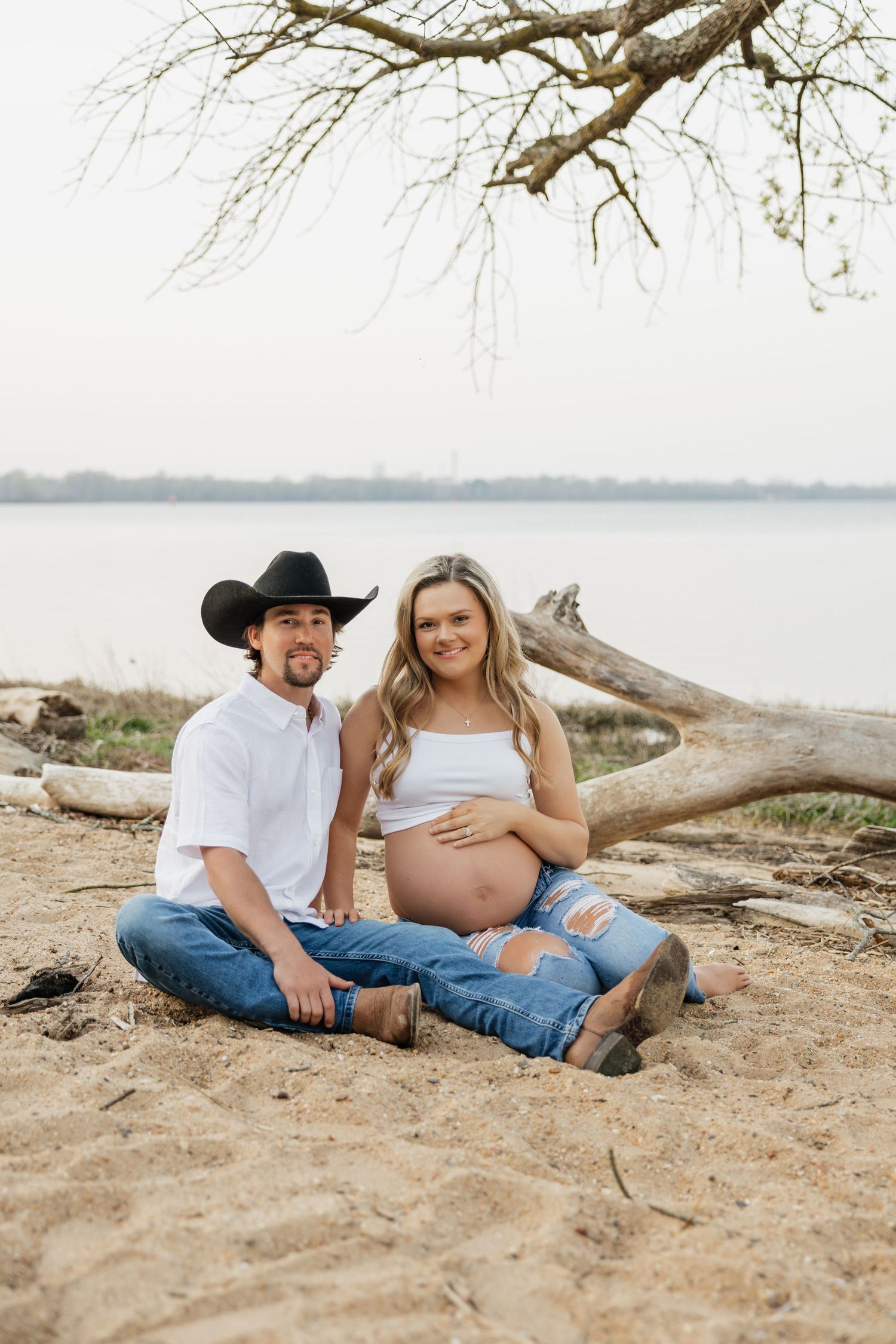Smiling couple sitting on sandy lakeshore, man in black cowboy hat beside pregnant woman in ripped jeans.