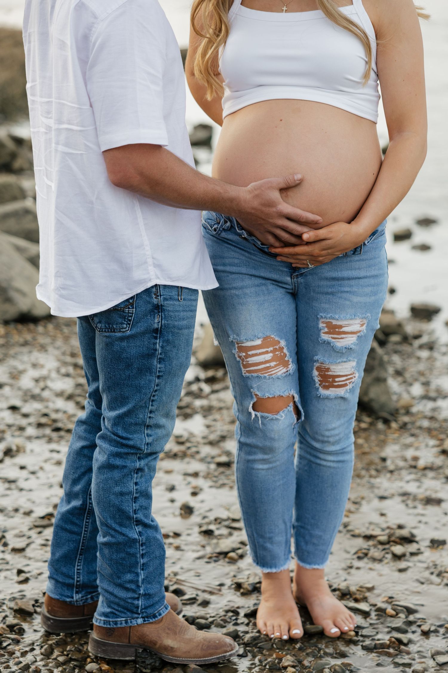Close-up of pregnant belly with couple holding baby bump on rocky beach, woman in ripped jeans.