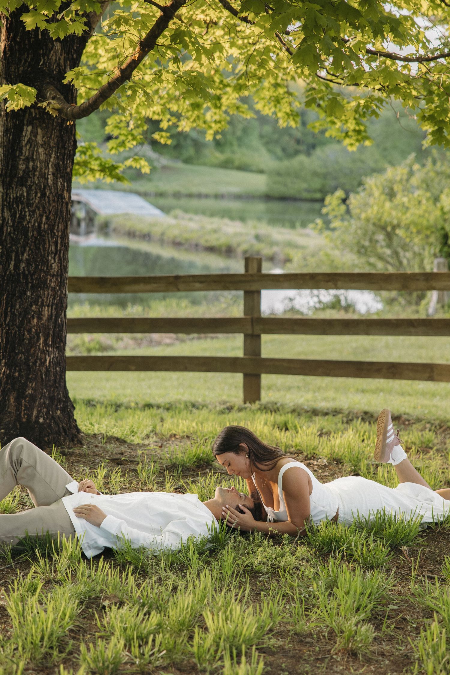 Couple relaxing on grass under a large tree near a wooden fence and pond in a scenic park setting.