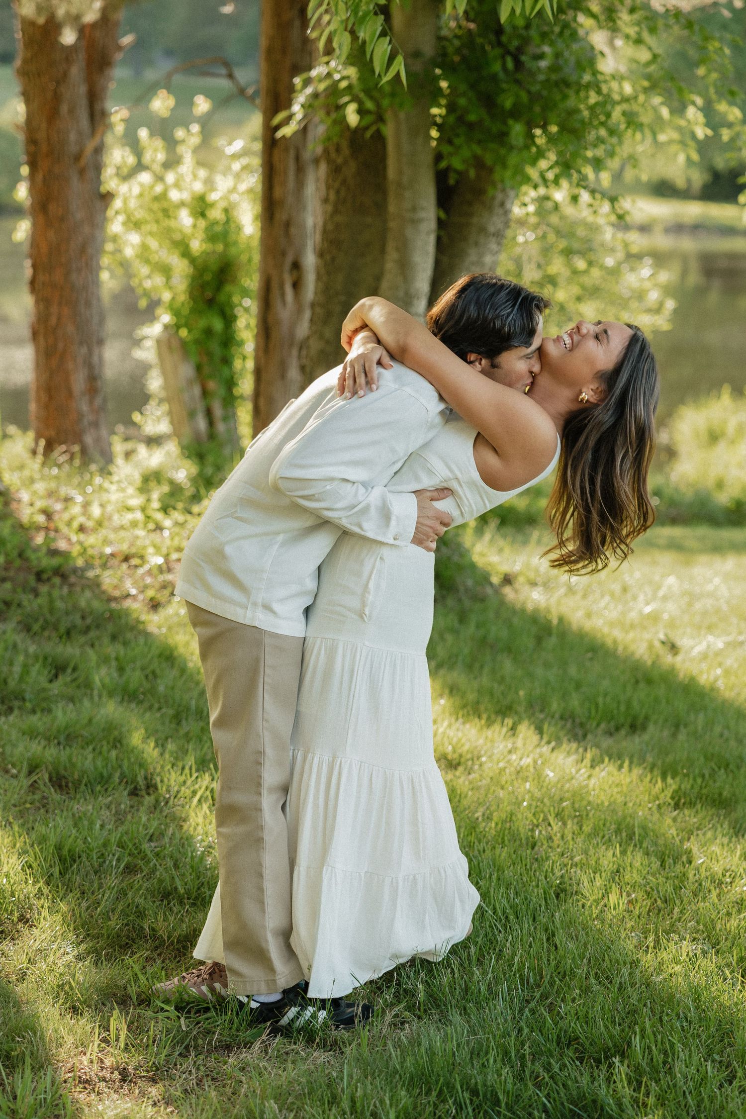 Couple sharing a tender kiss while embracing in a sun-dappled green garden near a peaceful lake.