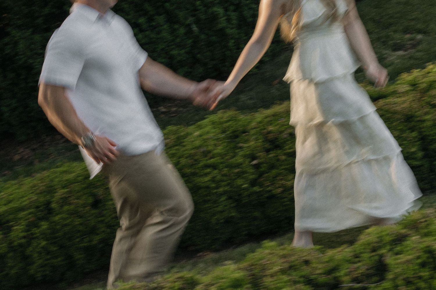 Blurred motion shot of couple holding hands running through lush green hedges in soft natural light.