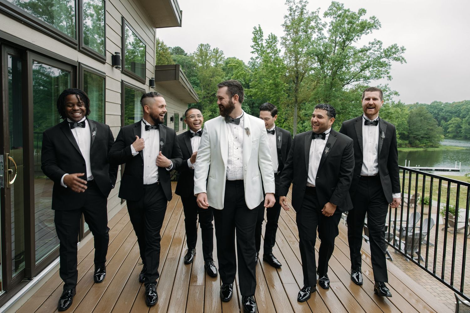 Groom in white tuxedo jacket laughs with groomsmen in black tuxedos on outdoor wooden deck.