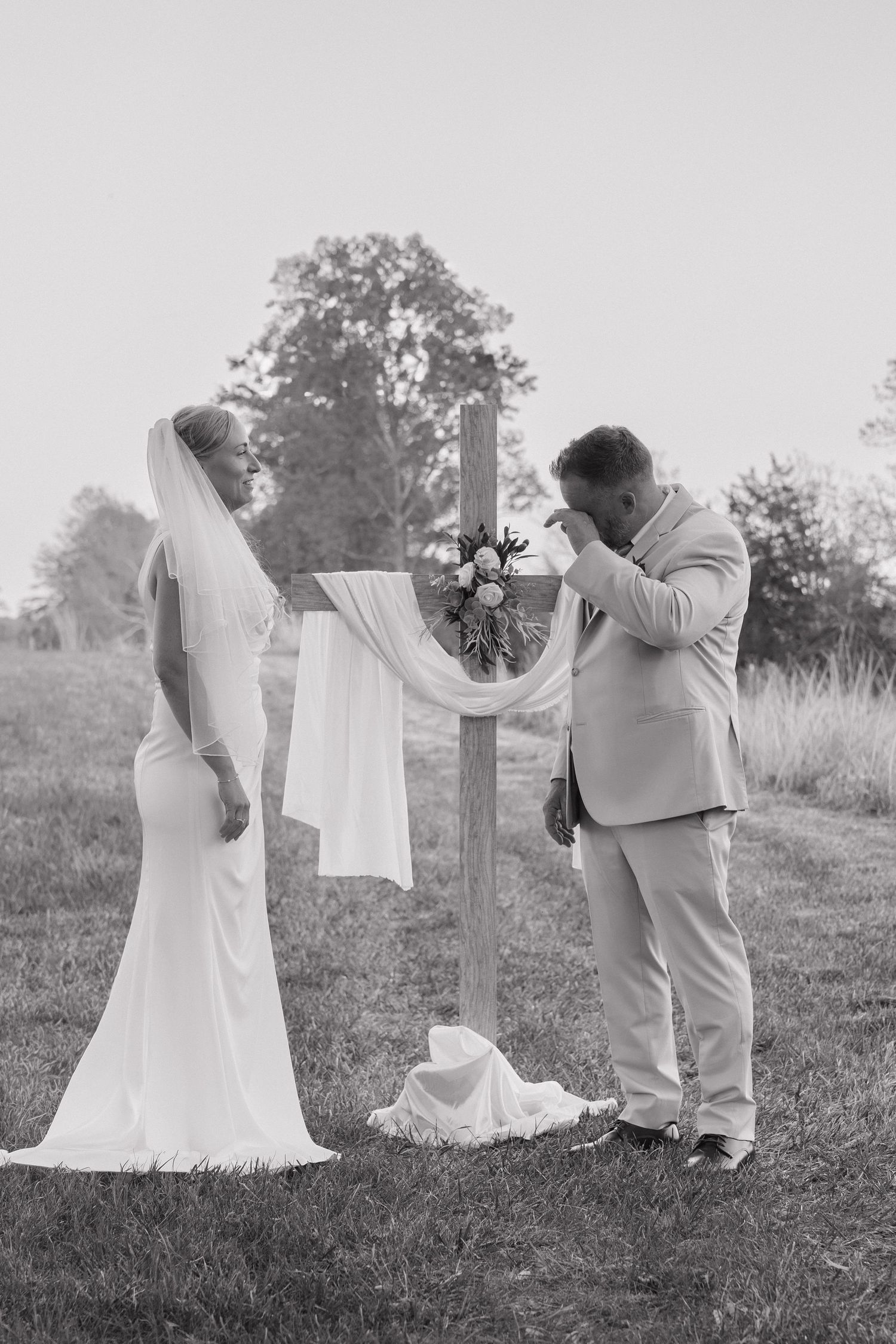 Groom wiping tears as bride approaches rustic wooden cross altar at outdoor wedding ceremony.