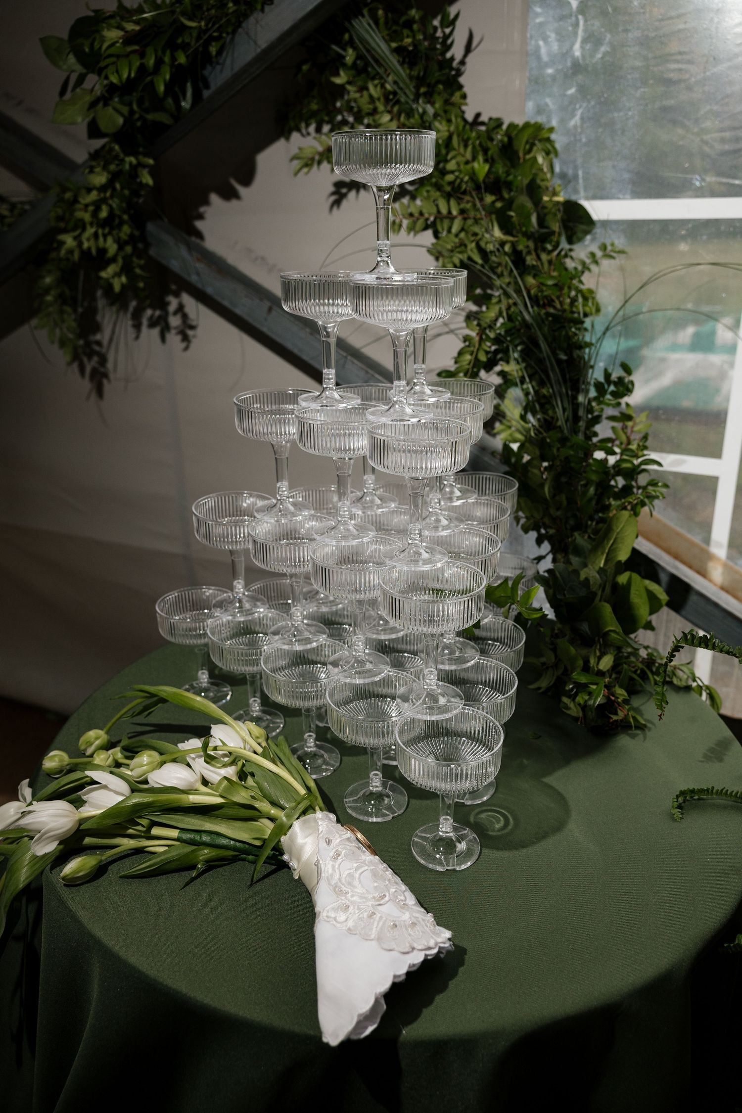 Elegant champagne coupe tower on green table with bridal bouquet and lush greenery floral arch.