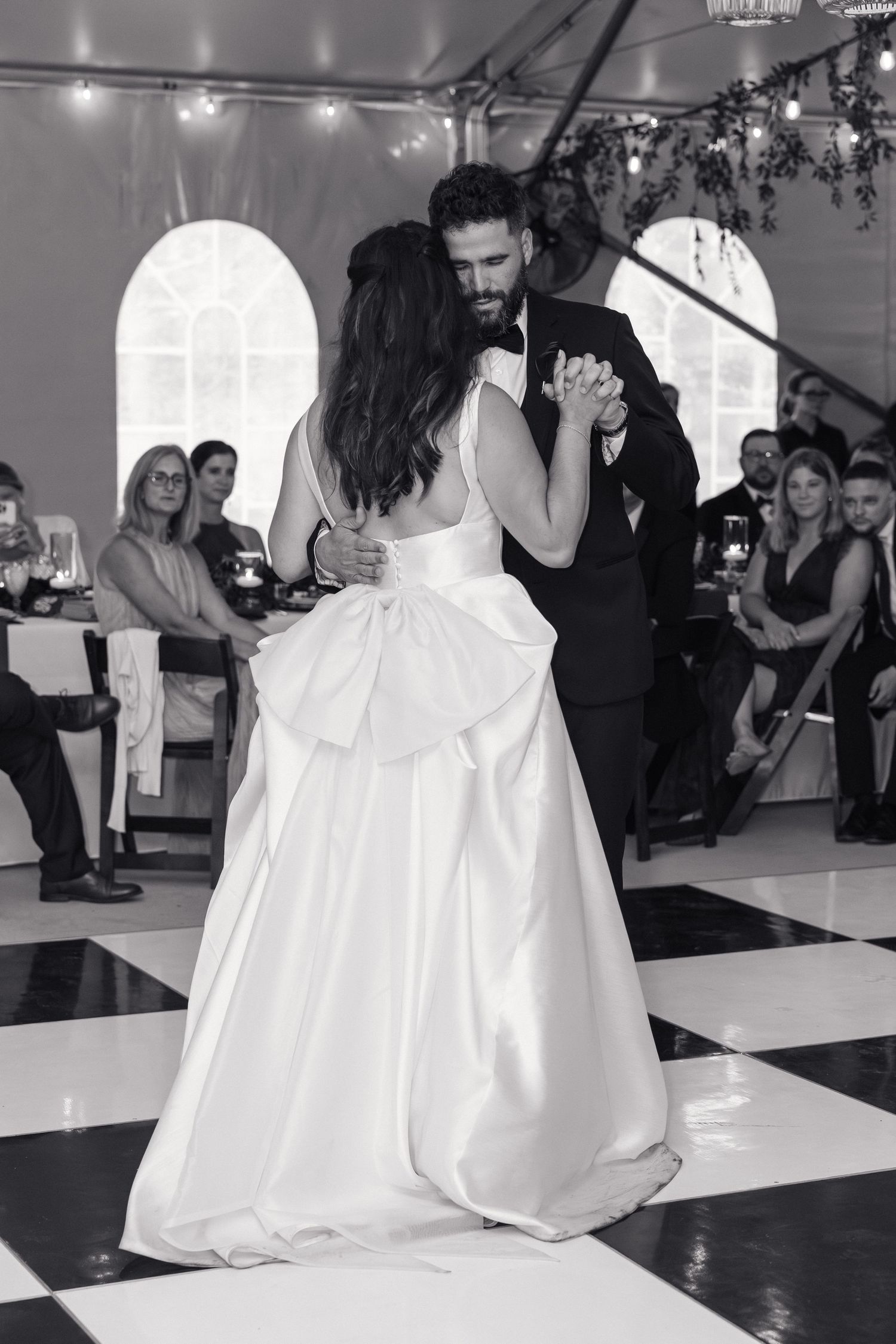 Black and white photo of bride and groom dancing closely on checkered floor before wedding guests.