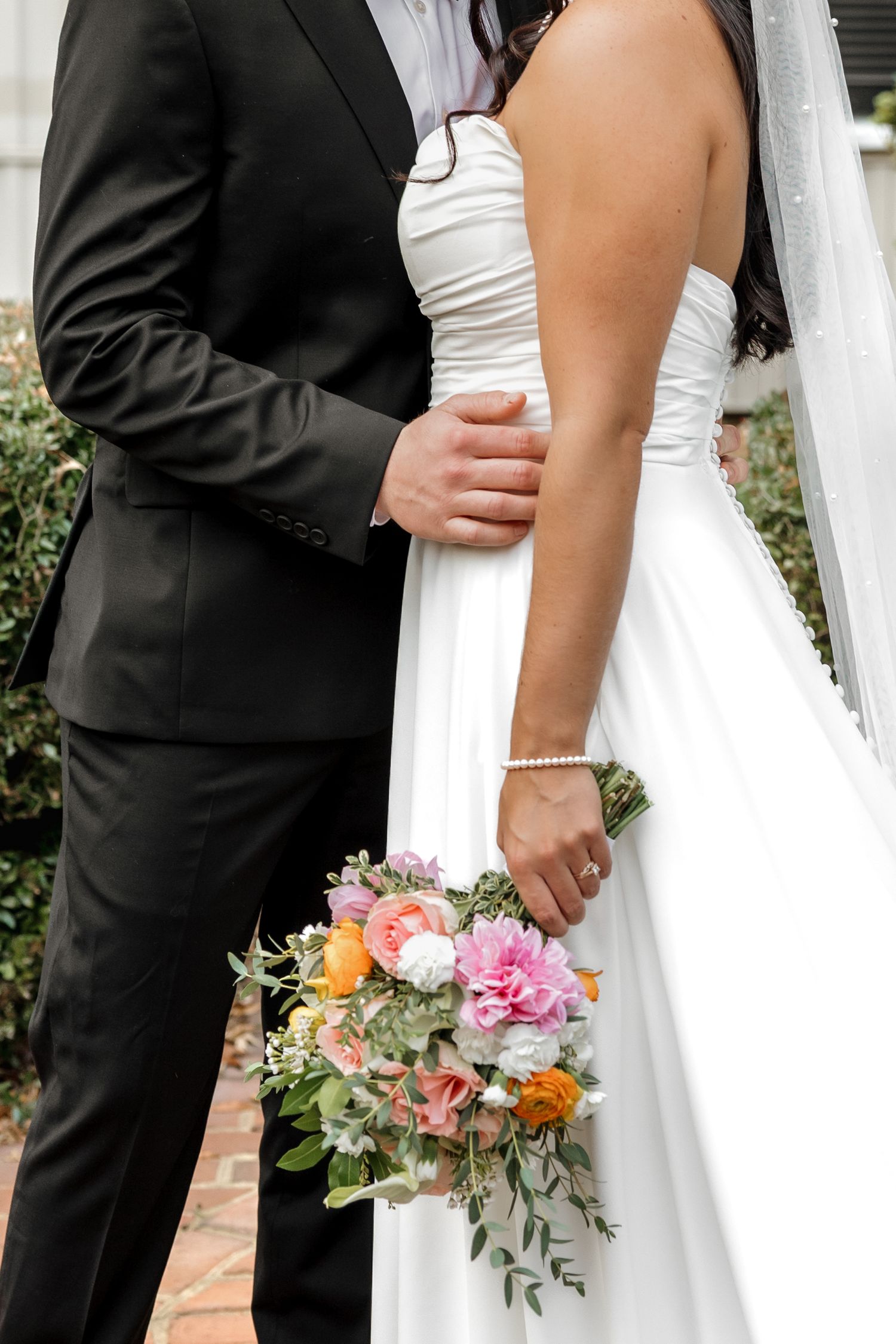 Close-up of bride holding colorful bouquet while groom places hand on her waist outdoors.