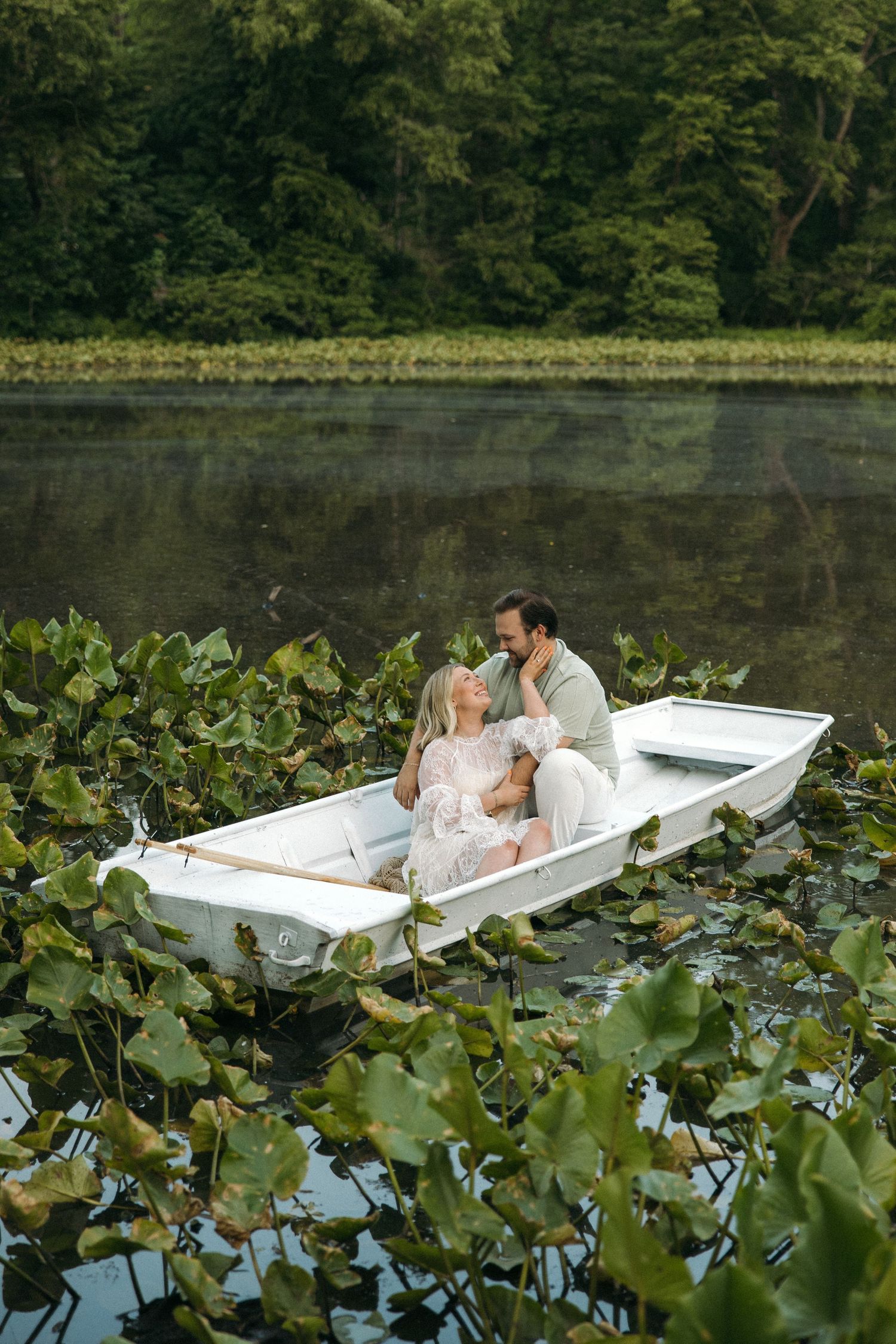 A romantic couple sits closely together in a white boat amid green lily pads on a tranquil forest lake.