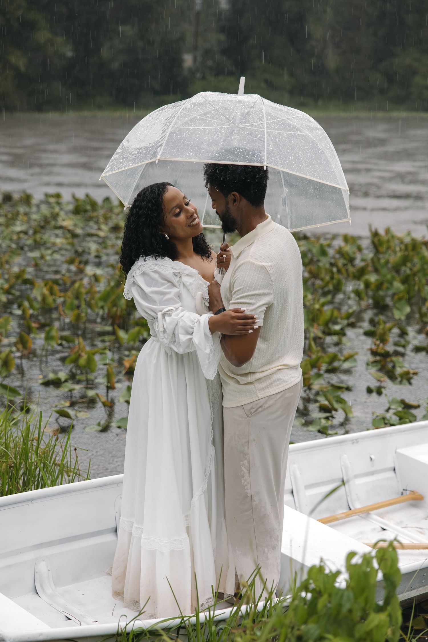A romantic Black couple in white clothing share a clear umbrella while standing in a white boat during rain.