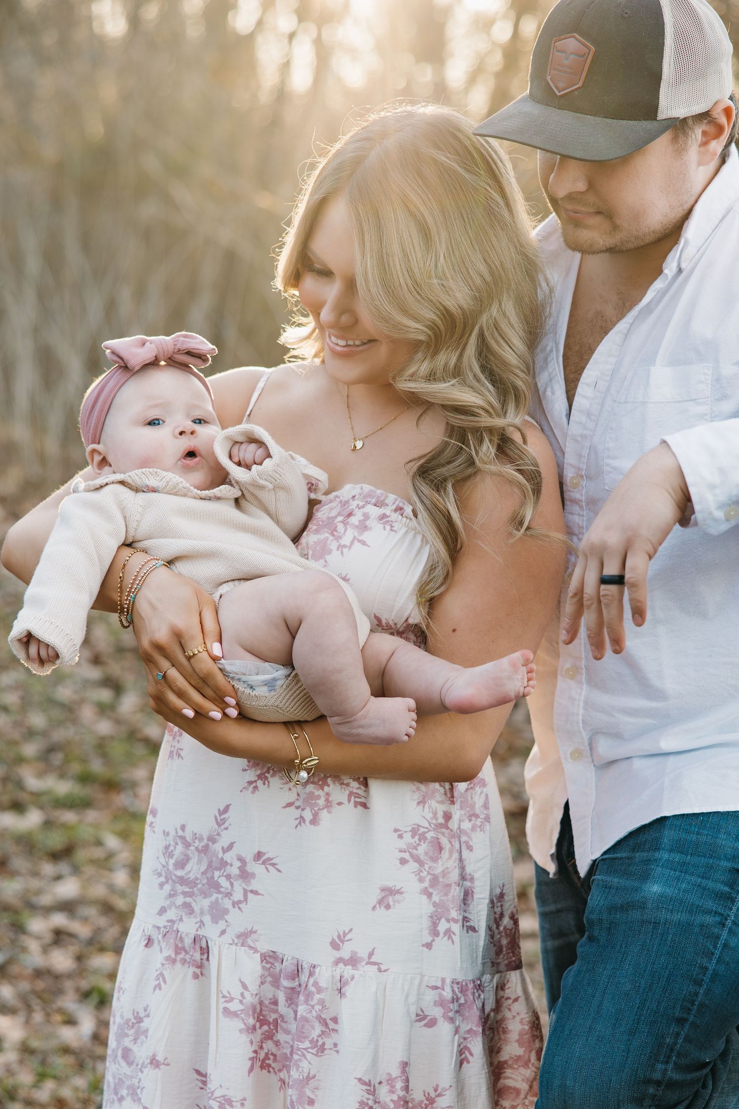 Young family with newborn baby at golden hour, mother in floral dress holding infant outdoors.