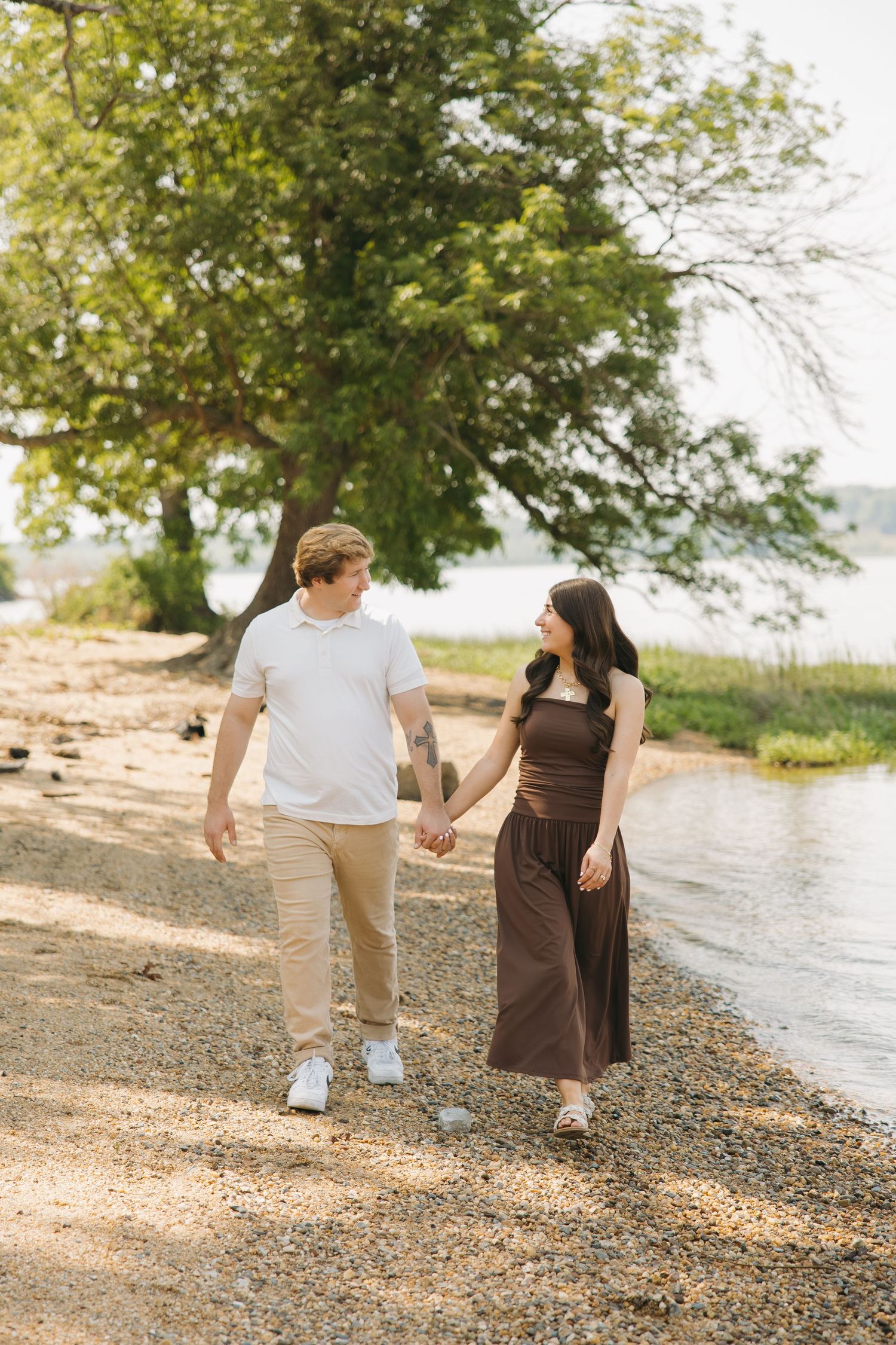 Engaged couple holding hands and walking along a rocky shoreline with trees and water in background.