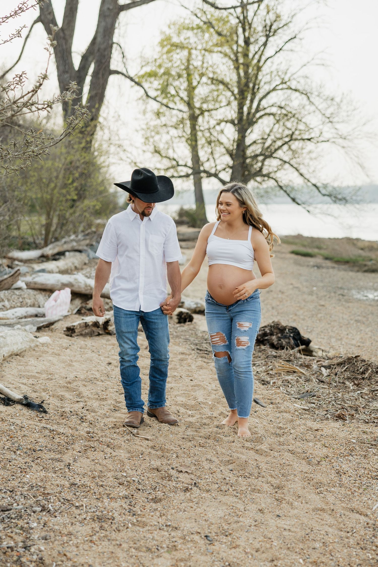 Smiling pregnant couple walks hand in hand on sandy beach near lake, man wearing black cowboy hat and boots.