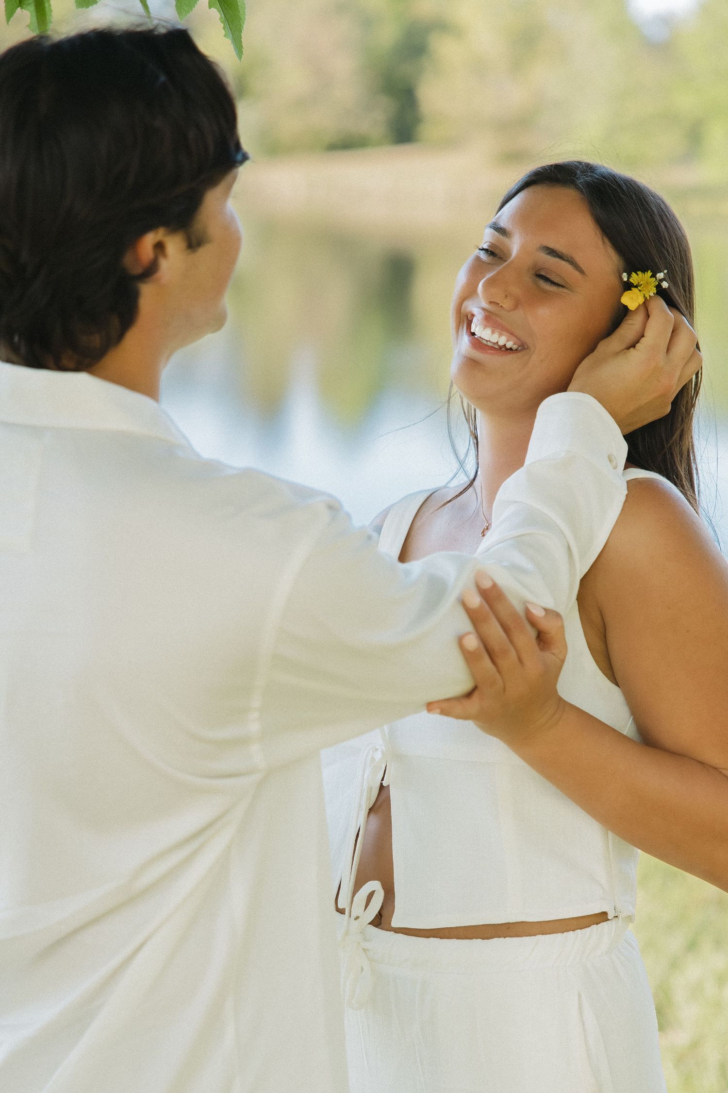 Man placing a yellow flower in laughing woman's hair near a sunlit lake, both dressed in white outfits.