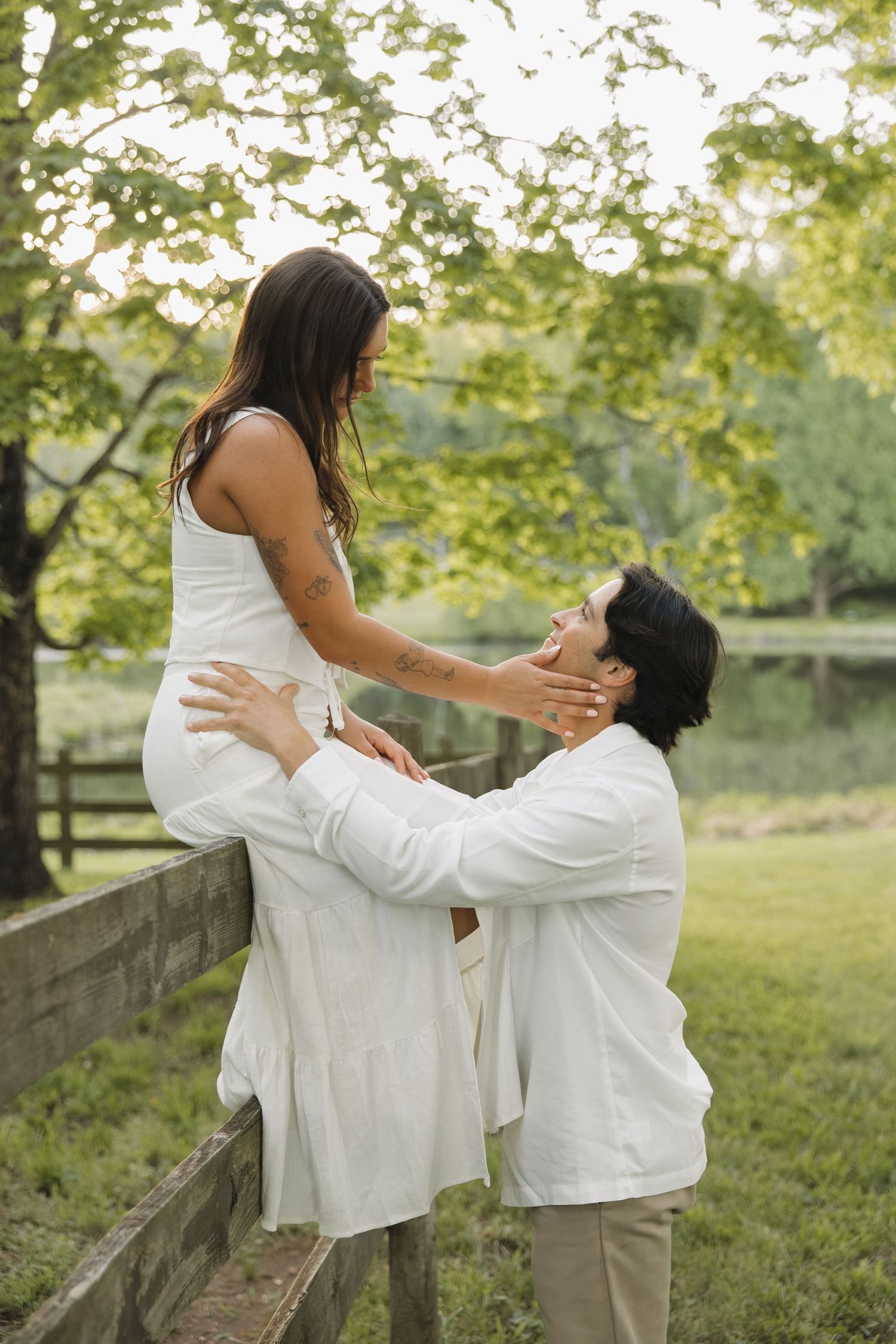 Man lifting woman onto a wooden fence while gazing at each other lovingly in a green park.