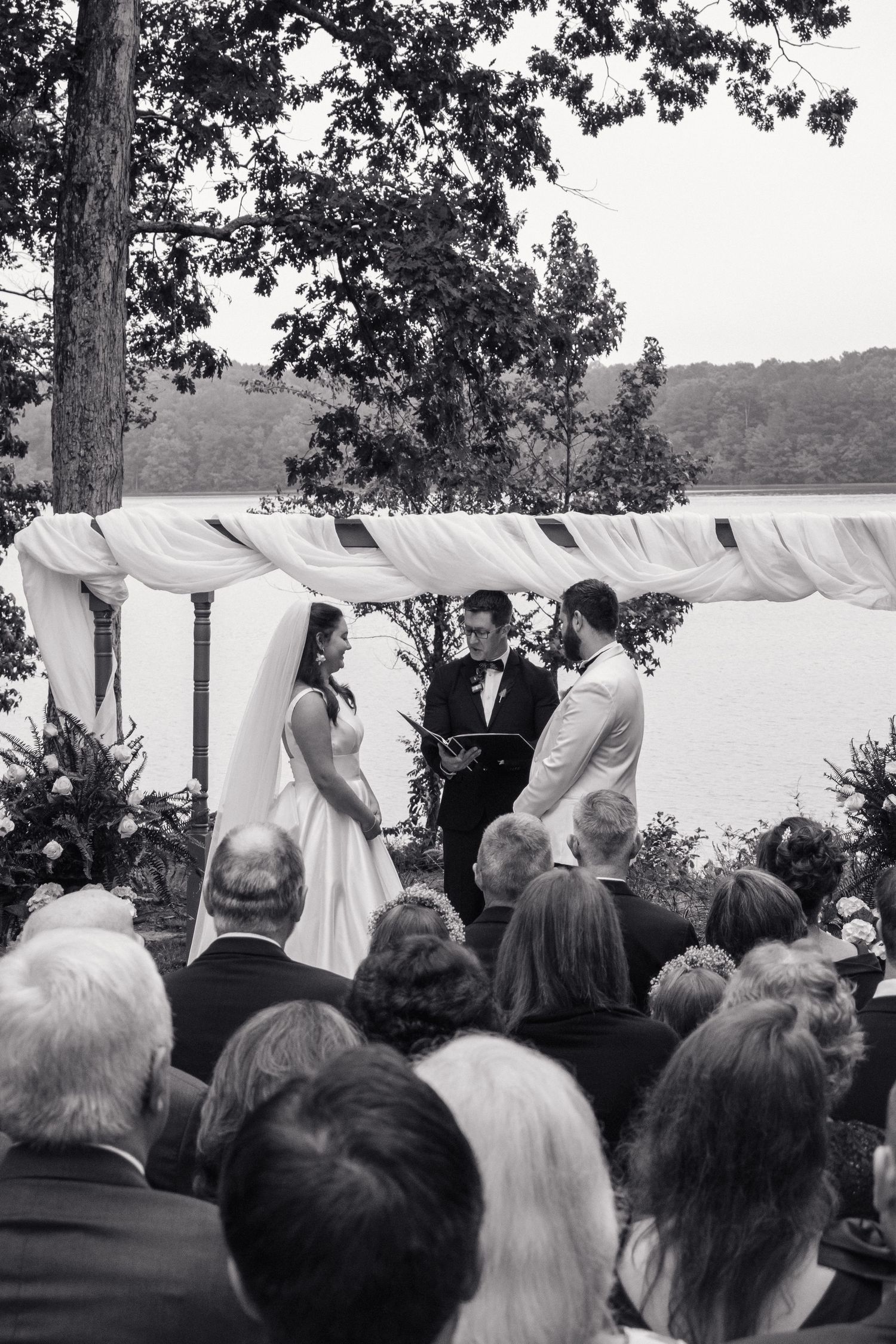 Black and white photo of bride and groom exchanging vows under draped arch near lakeside with seated guests.