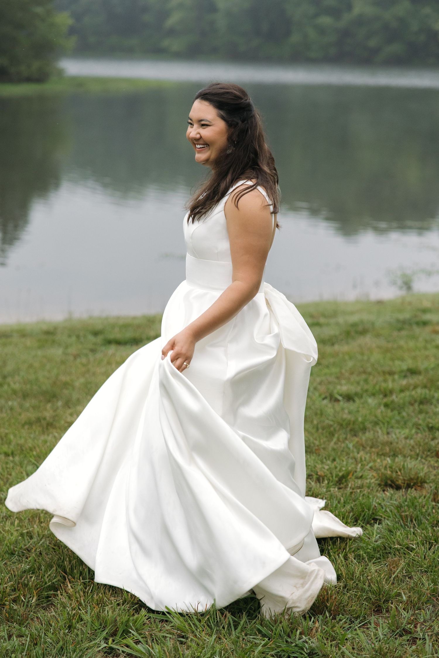 Radiant bride in white satin gown twirling beside a serene misty lake.