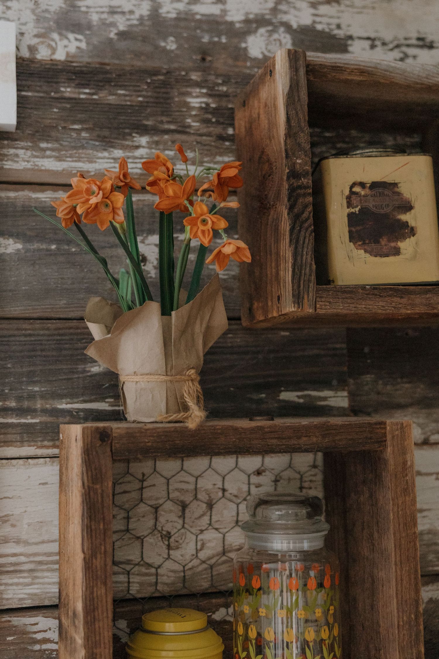 Orange flowers in kraft paper wrap on rustic reclaimed wood shelving with vintage decor.