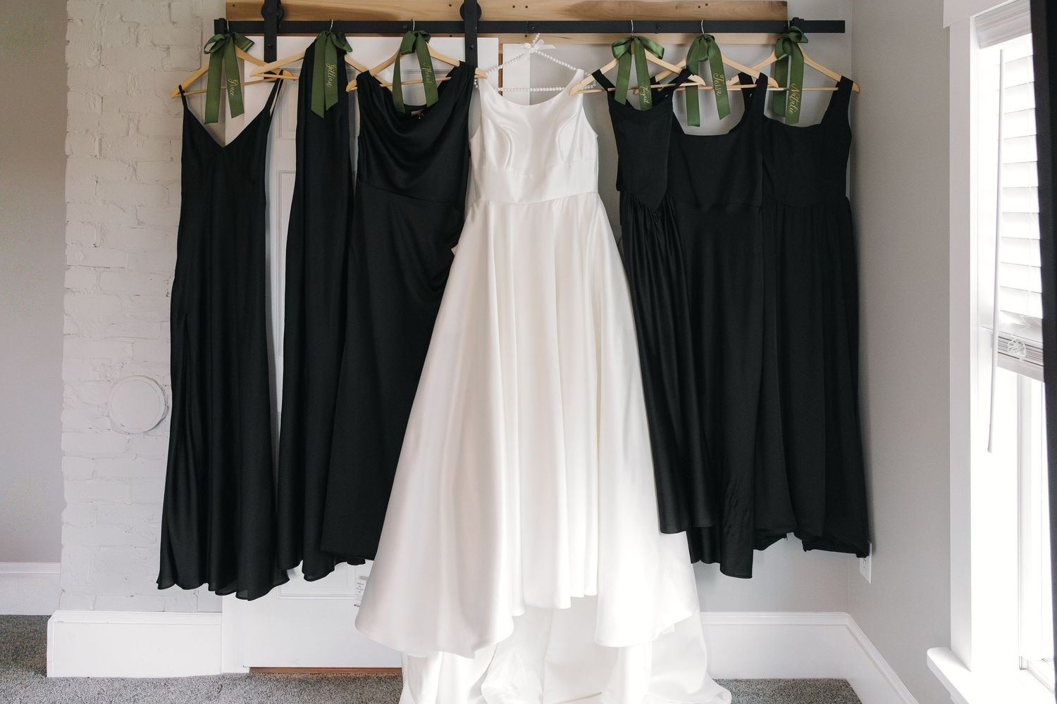 White bridal gown centered among four black bridesmaid dresses hanging on a wooden rack with green ribbon hangers.