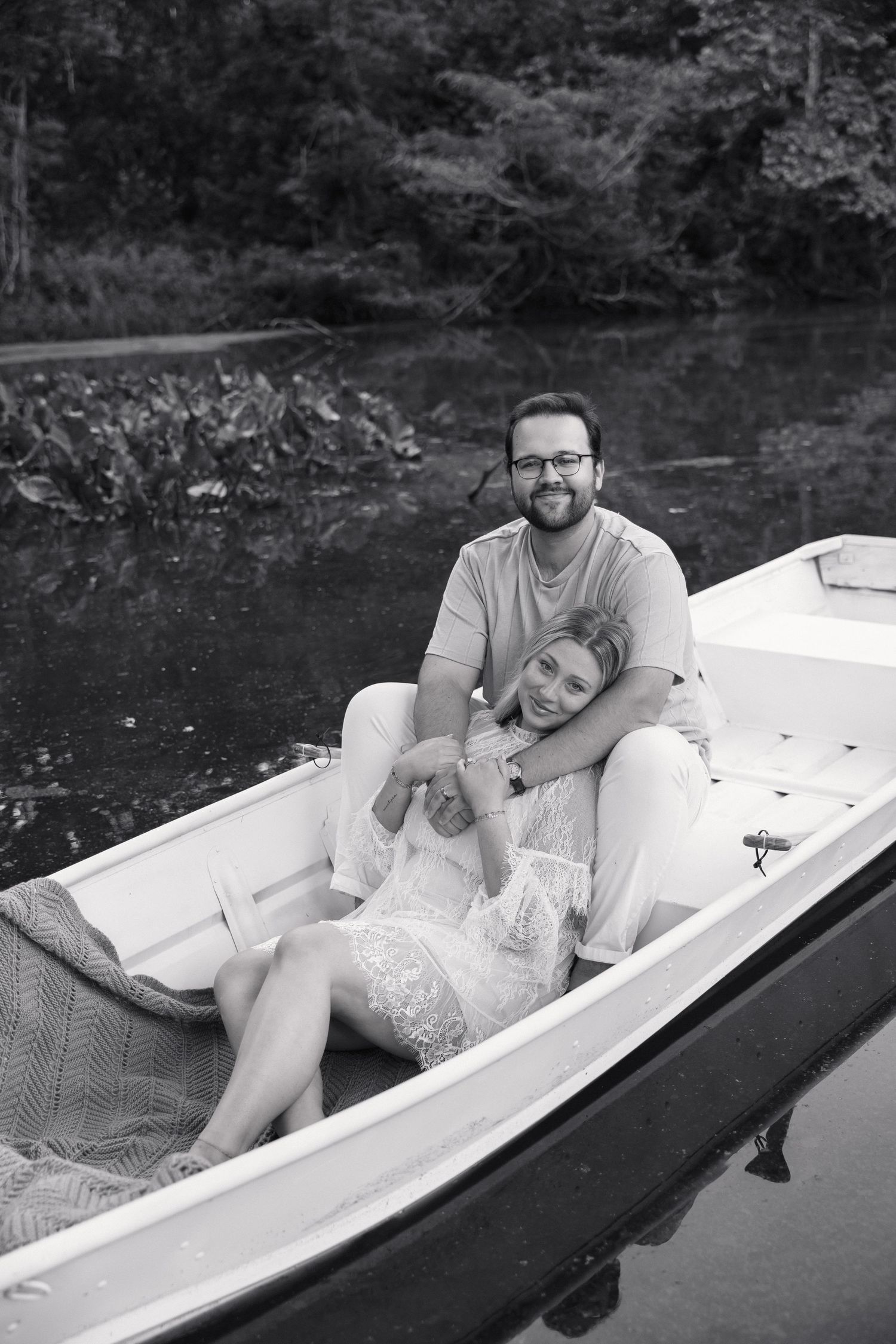 Black and white portrait of a couple cuddling in a white rowboat on a tranquil river.