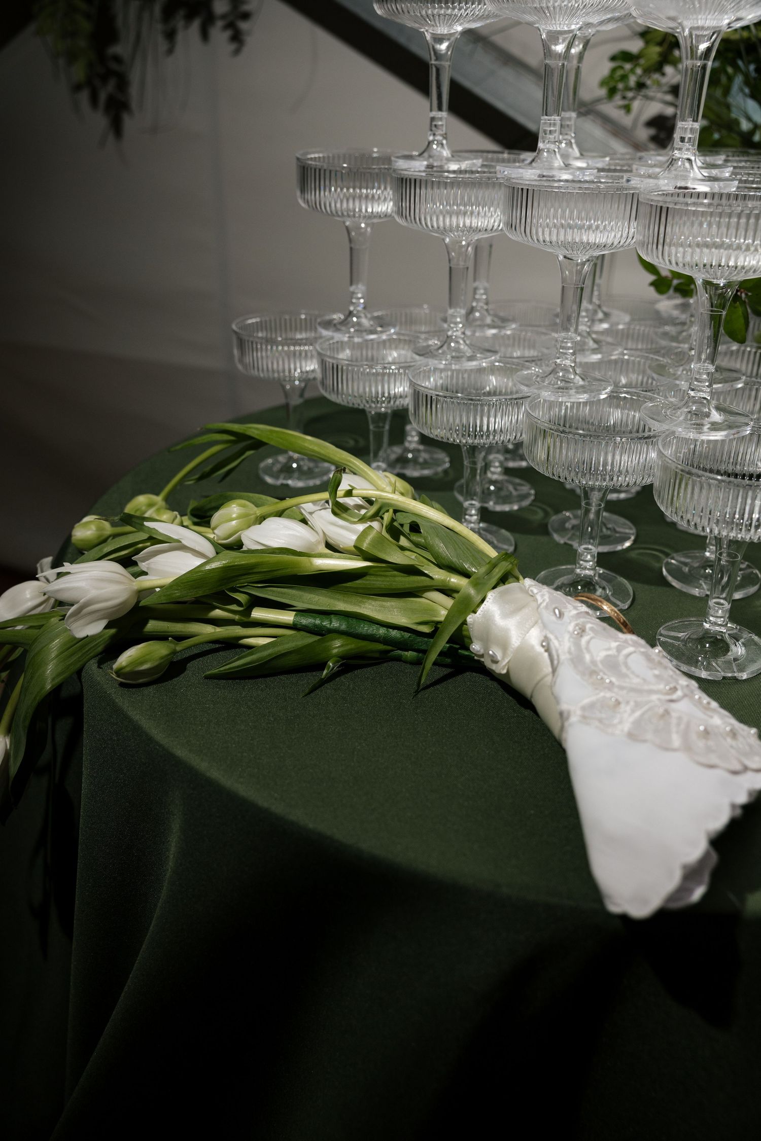 White tulip bridal bouquet beside champagne coupe glass tower on dark green tablecloth.