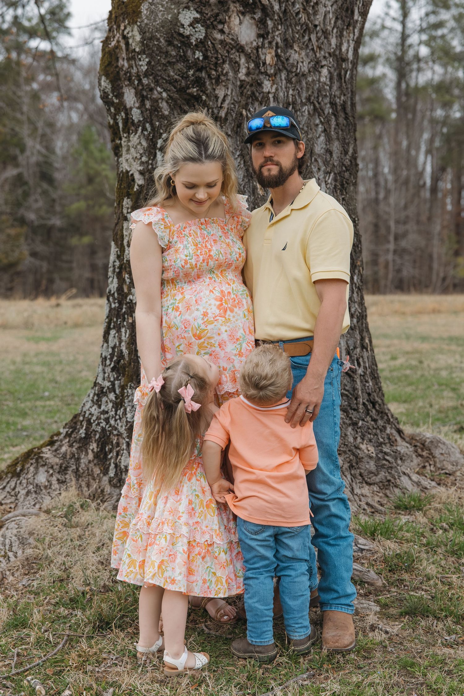 Pregnant mother and bearded father stand by a large tree as two young children hug them from behind.