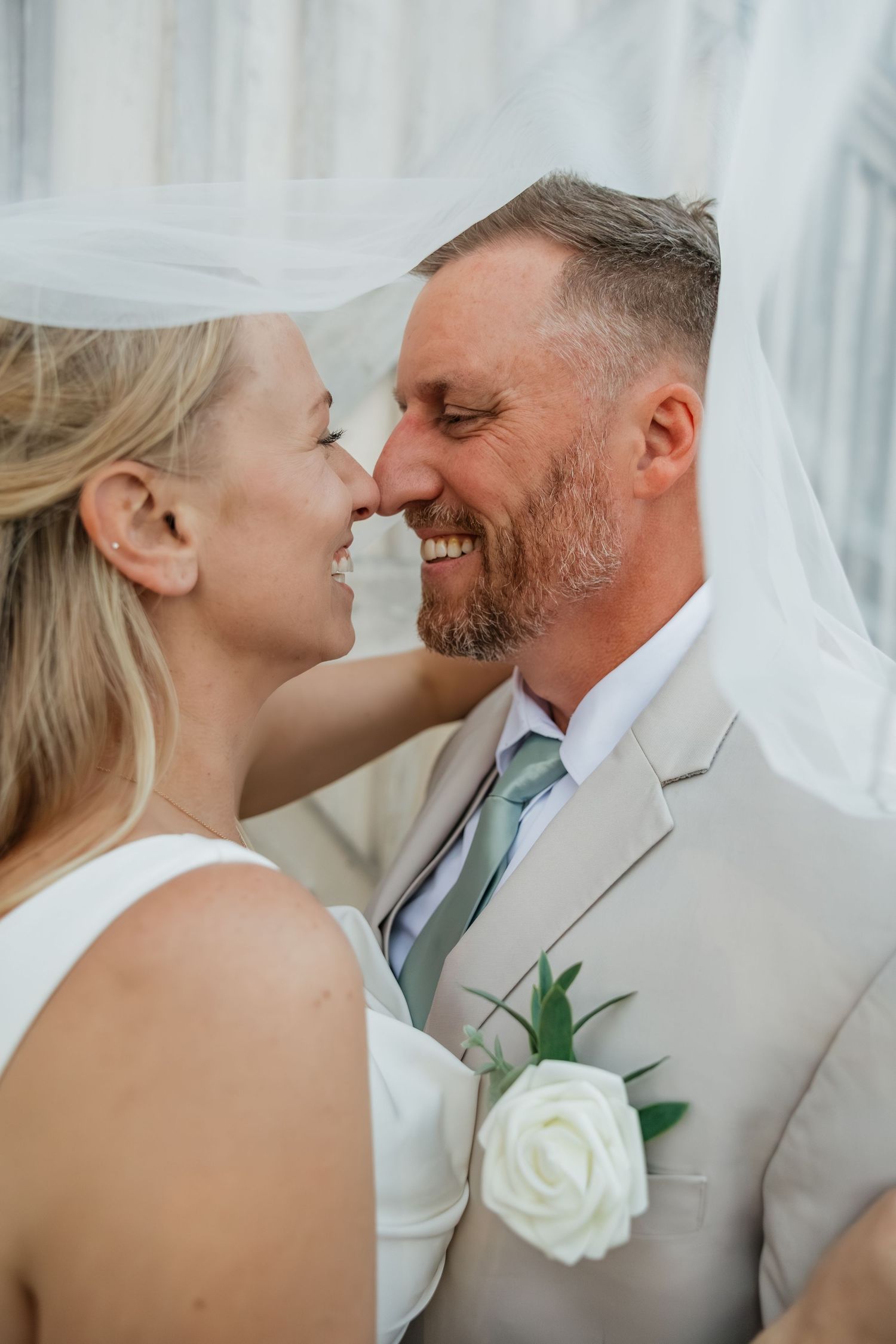 Smiling bride and groom share an intimate nose-to-nose moment under a flowing white wedding veil.