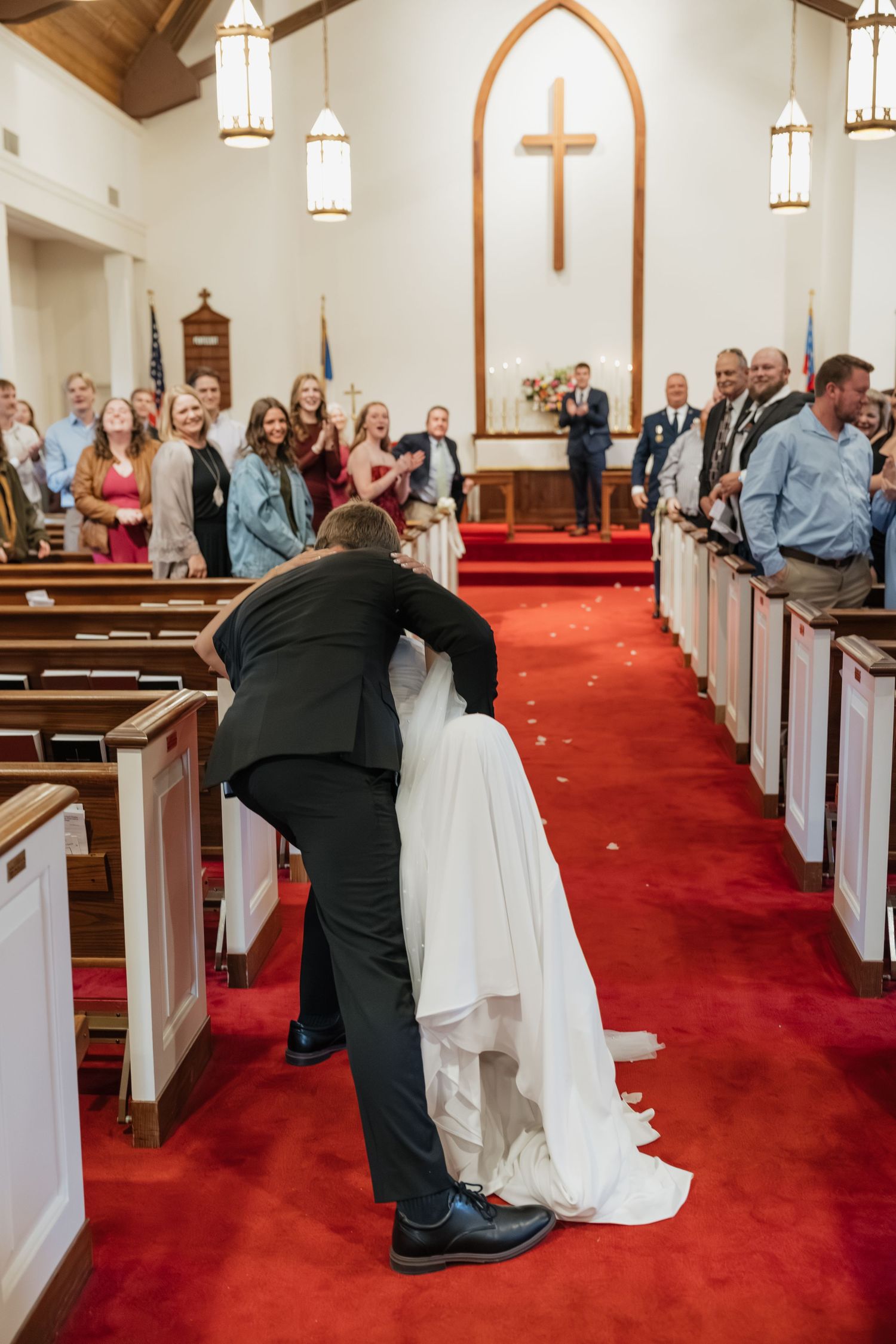 Groom dipping bride for a kiss in church aisle while guests cheer after wedding ceremony.