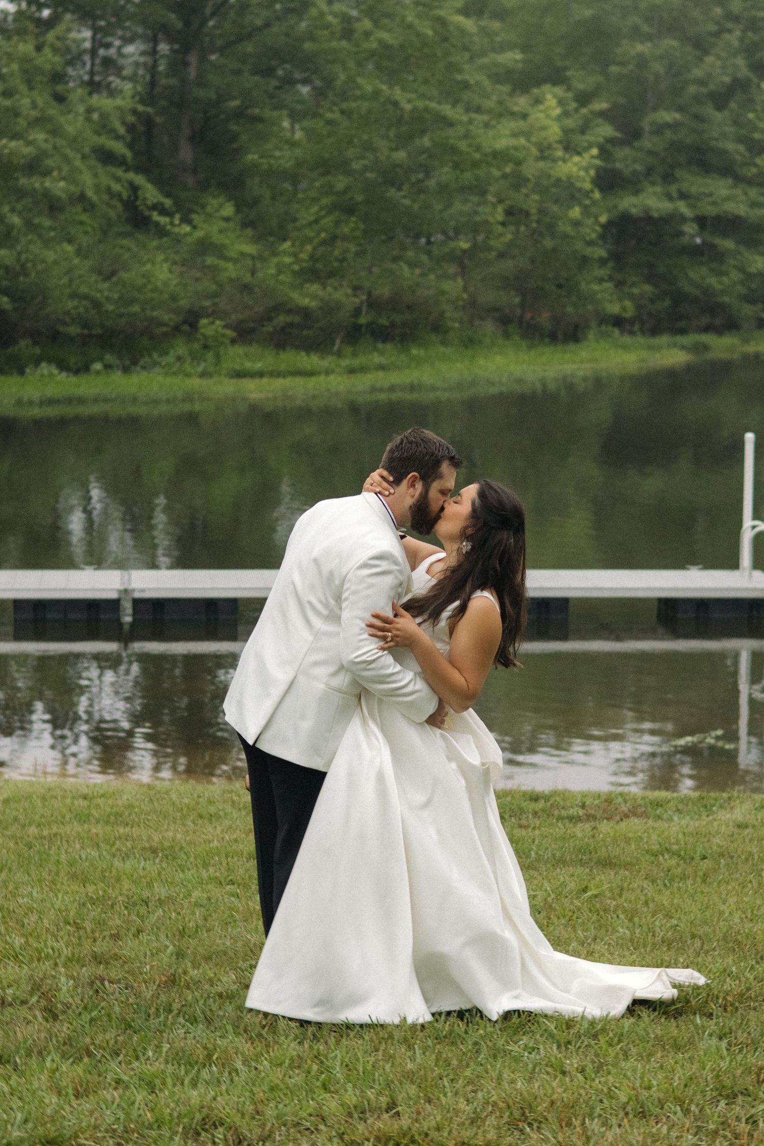 Bride and groom share romantic kiss beside calm lake surrounded by lush trees.