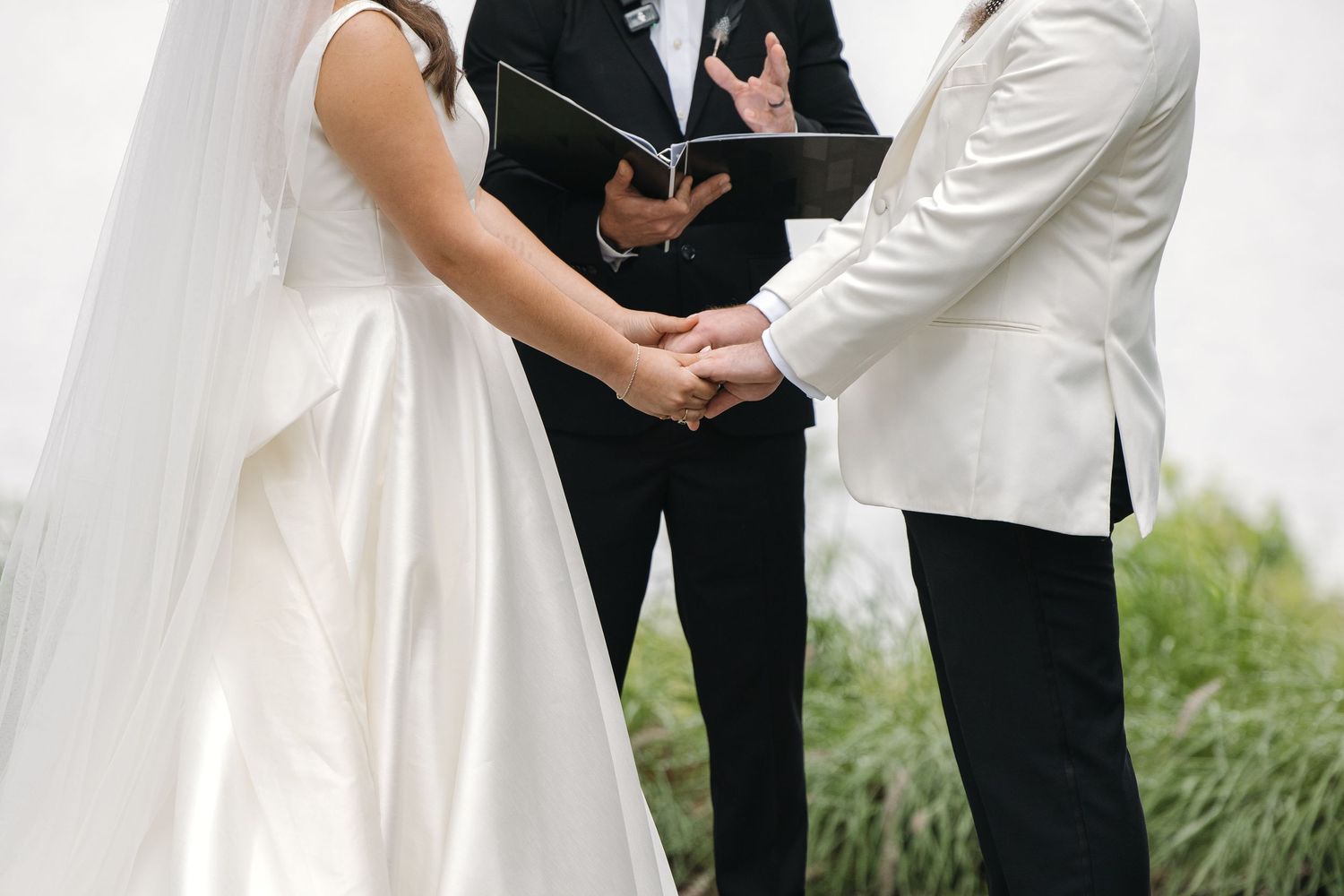 Bride and groom hold hands during outdoor wedding ceremony as officiant reads from a book between them.