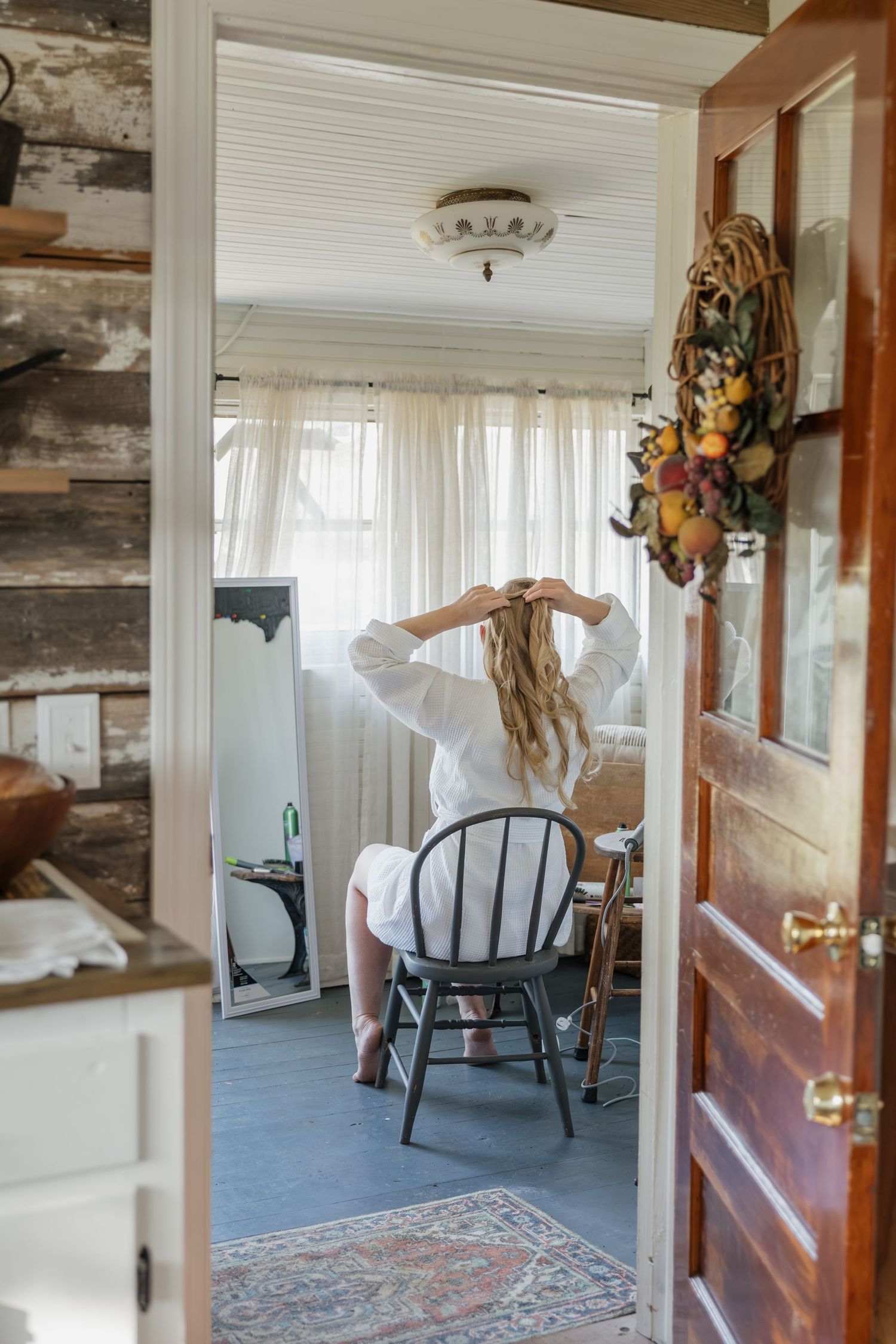 Bride in white robe doing hair in rustic log cabin room viewed through open doorway.