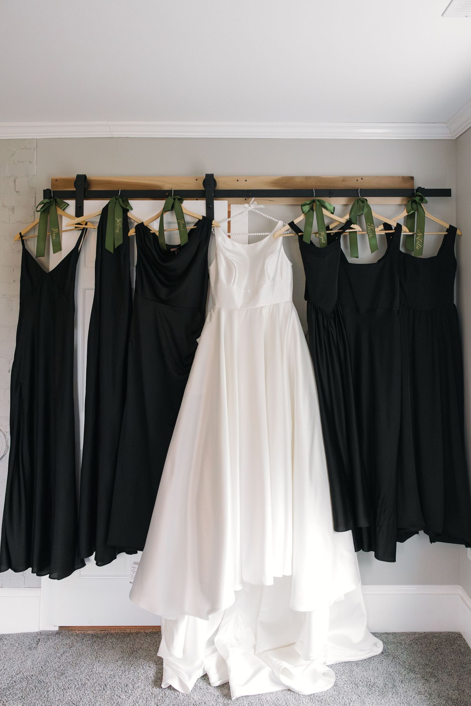White wedding dress flanked by four black bridesmaid gowns hanging on a wooden rack with green satin ribbon hangers.
