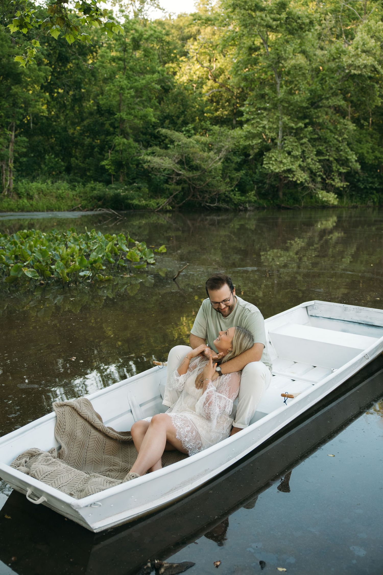 Romantic couple cuddling in a white boat on a still river with green forest backdrop.